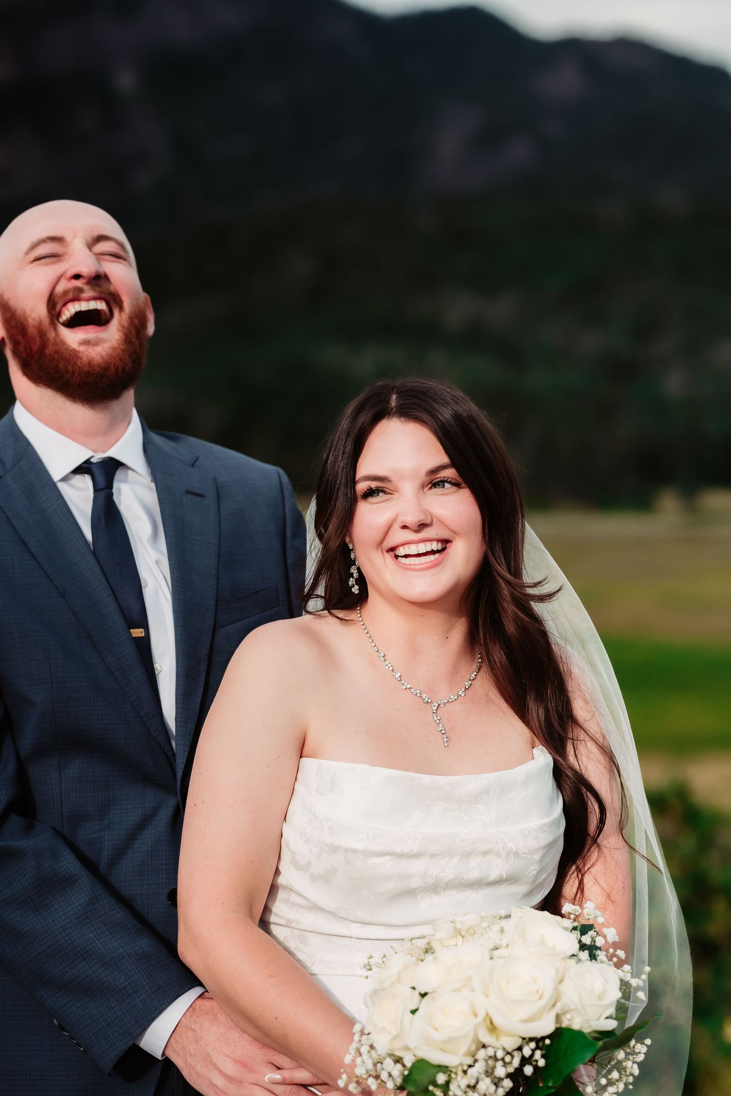 A joyful wedding couple laughing together in front of mountain scenery.