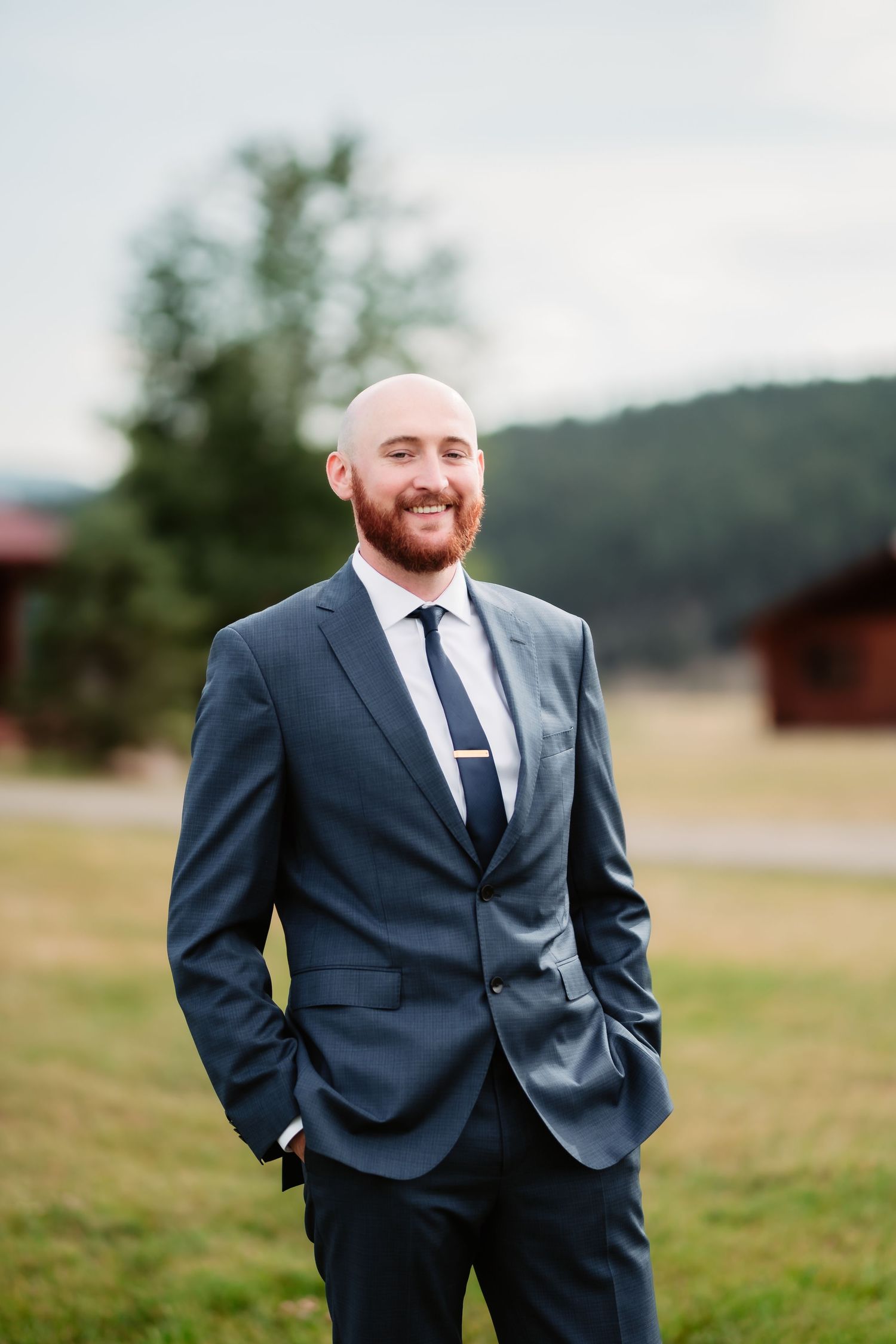 Groom in navy suit smiling outdoors with mountains in background.