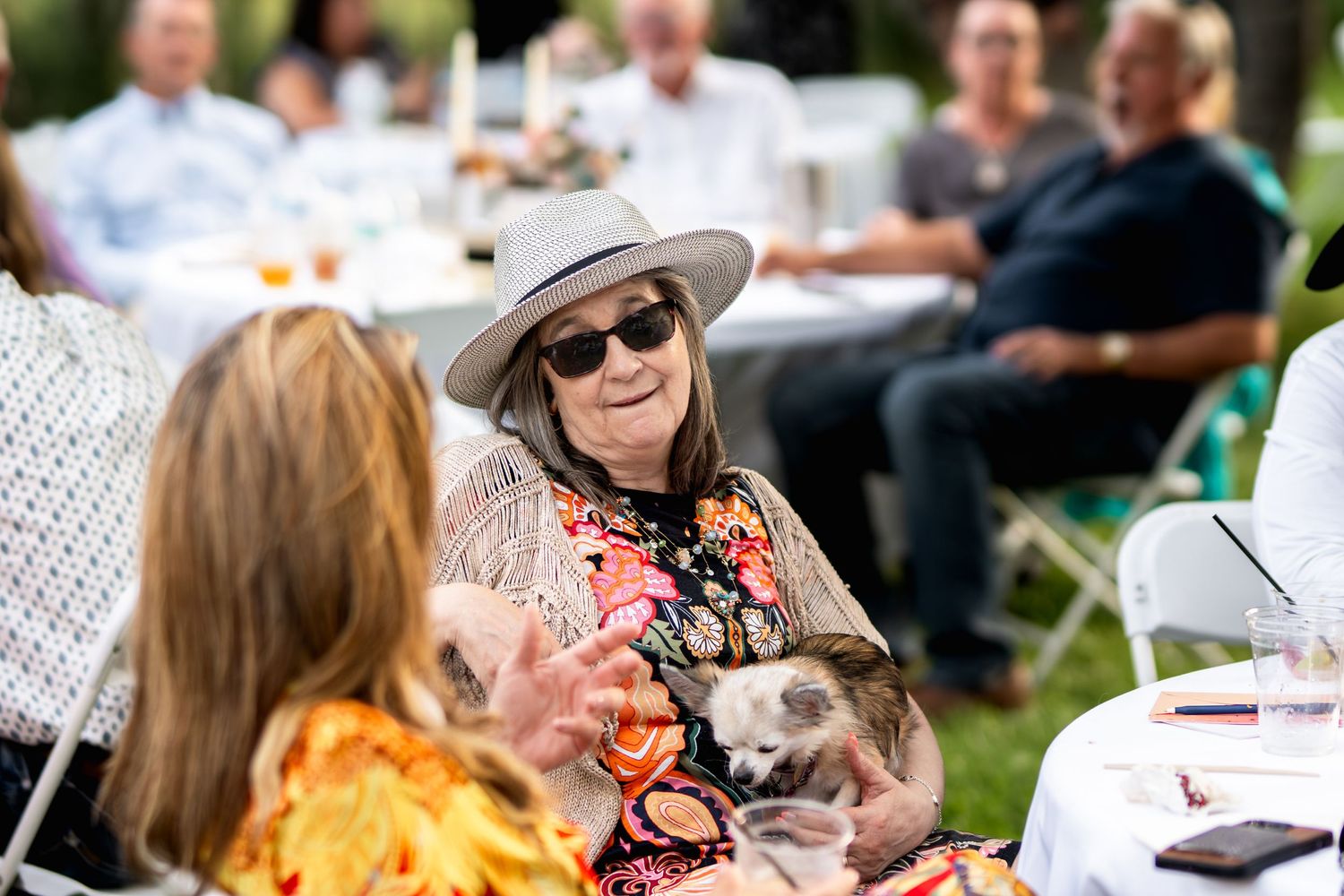 Wedding guest in sunhat and floral dress enjoying outdoor reception conversation.