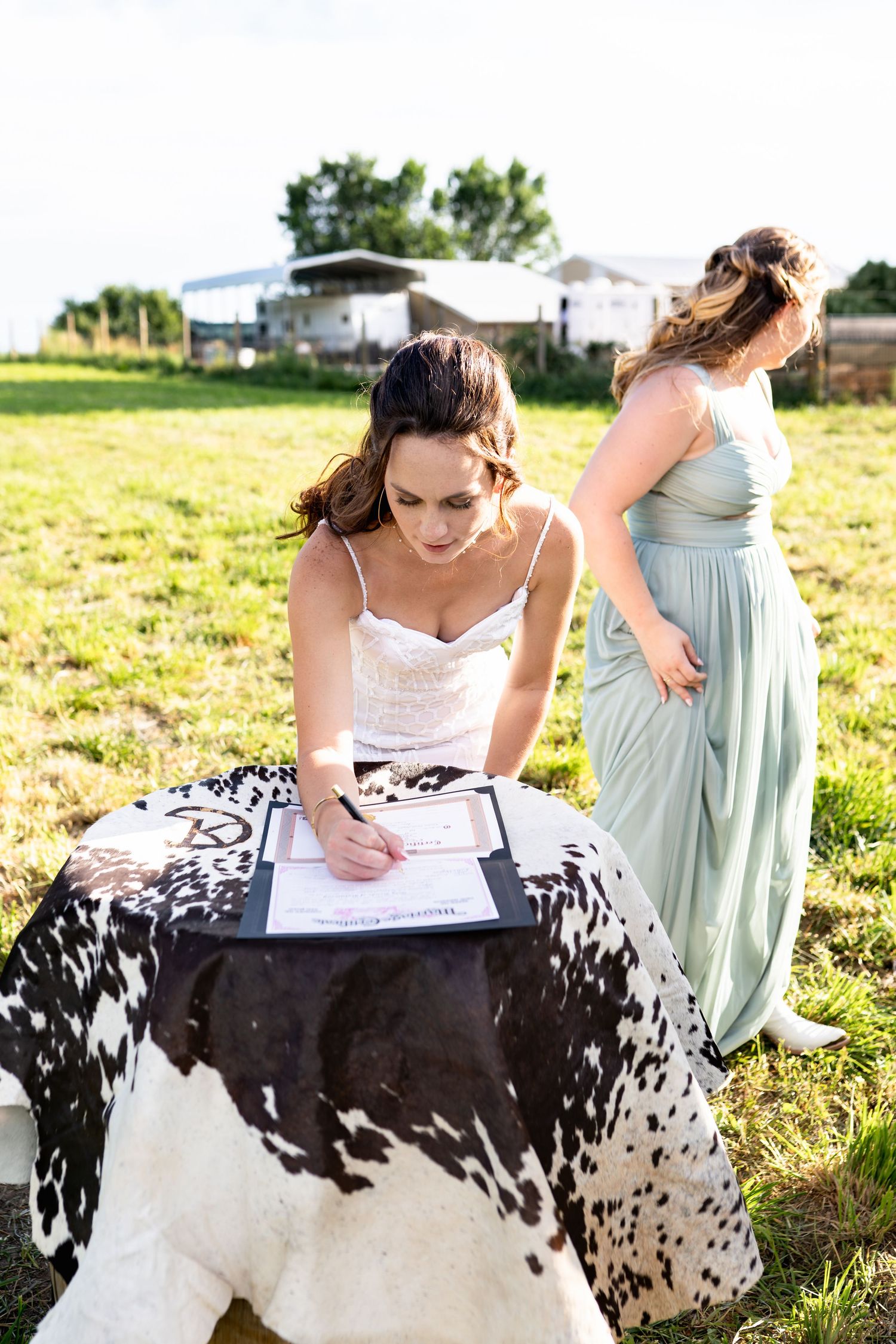Wedding guest in pastel dress signs a document on black and white cowhide covered table outdoors.