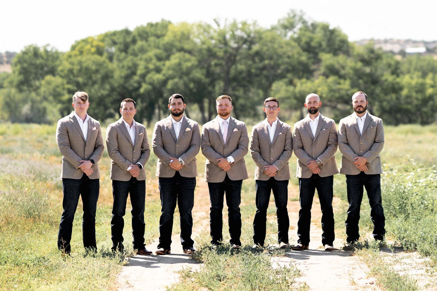 Wedding party groomsmen standing in a line wearing matching tan vests and suits in a sunlit outdoor field.