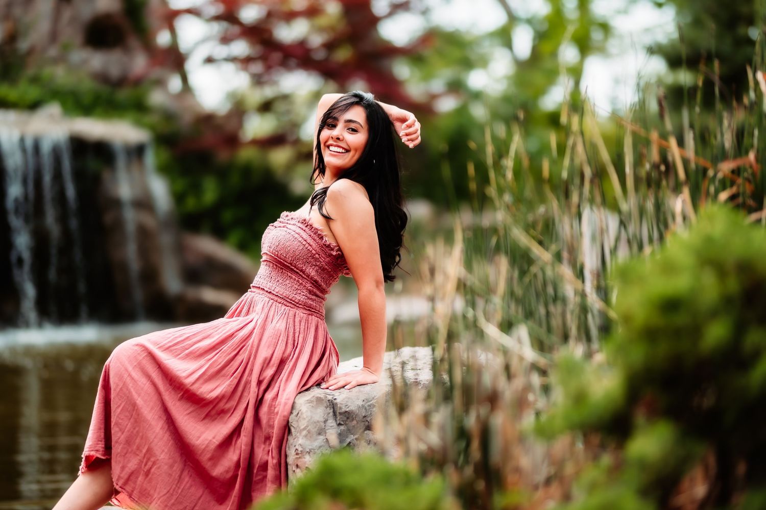 A person in a flowing pink dress poses gracefully by a waterfall in a lush garden setting.