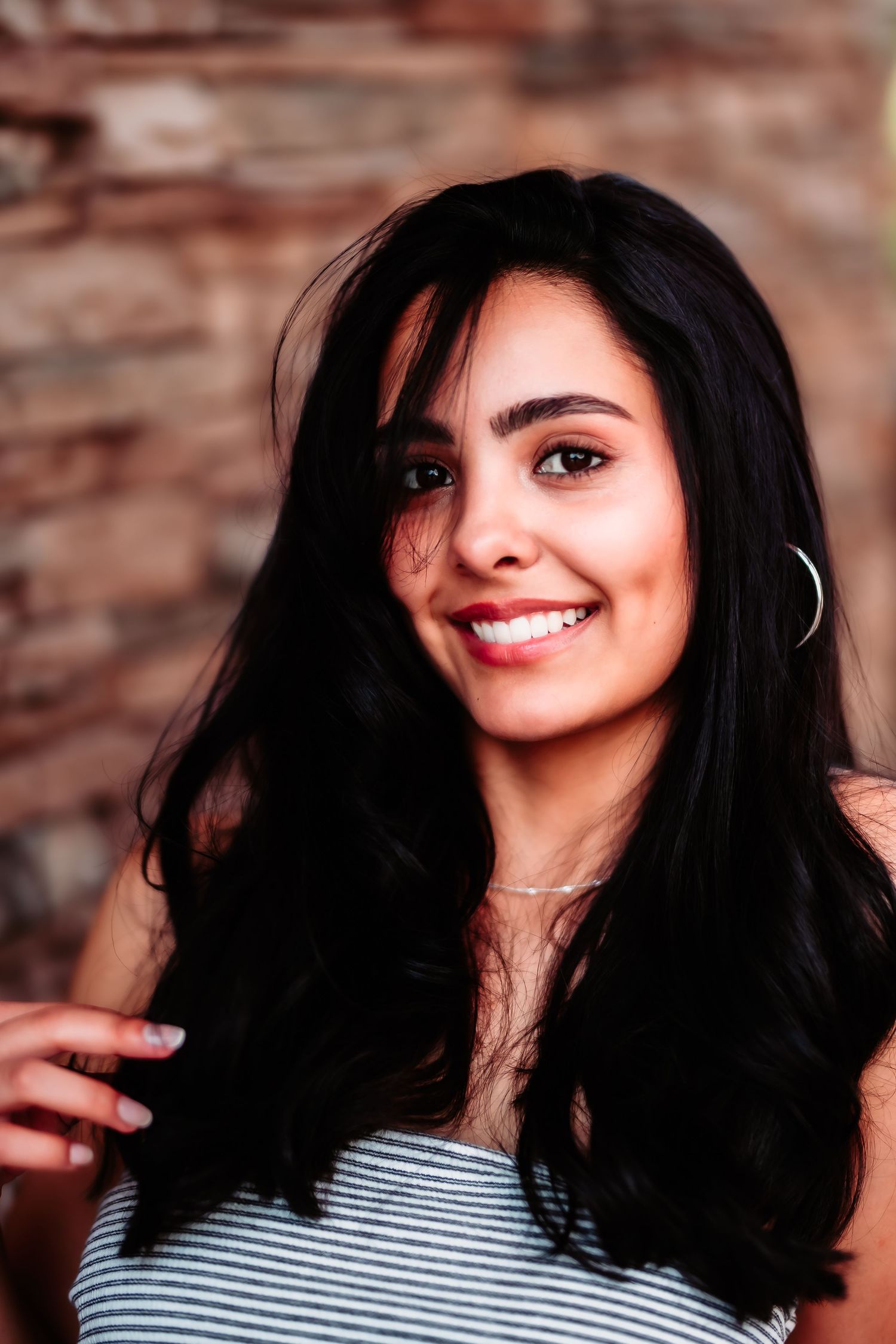 Portrait against a rustic brick wall showcasing long dark hair and a striped top in warm lighting.