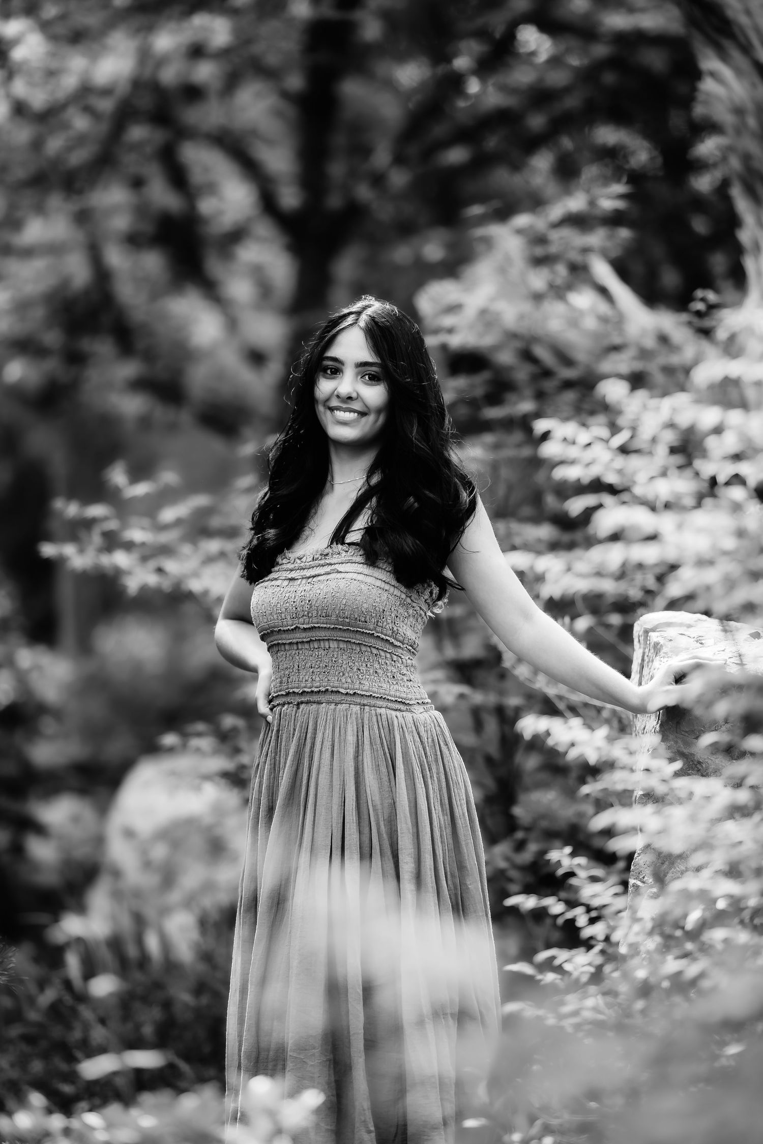 Black and white portrait of person in flowing dress standing among garden foliage.