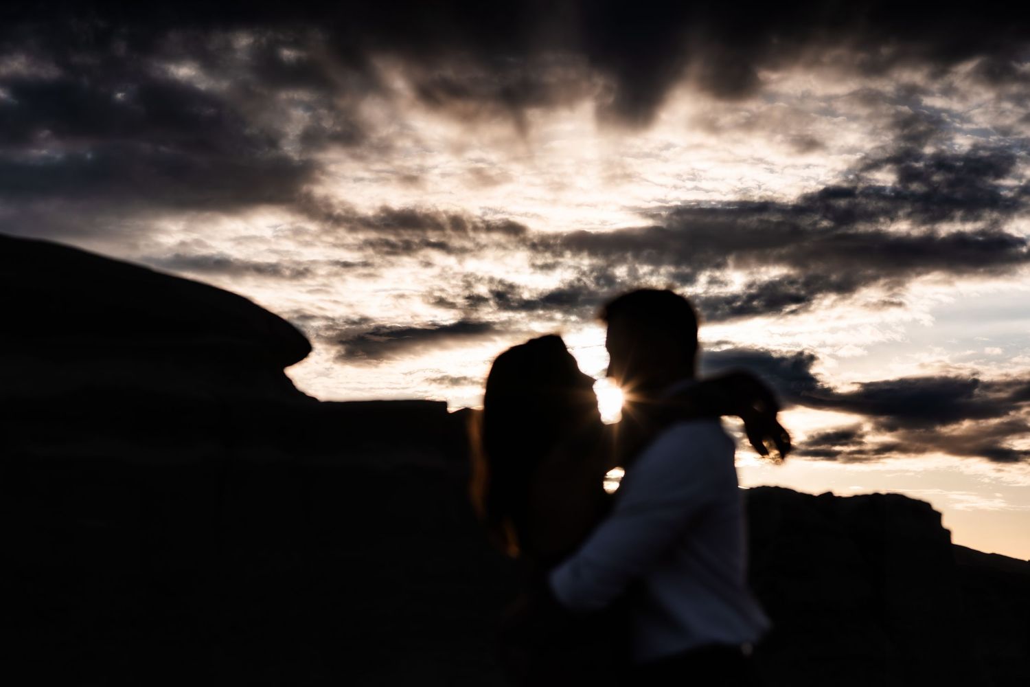 A silhouetted couple sharing an intimate moment against a dramatic sunset sky with rock formations.