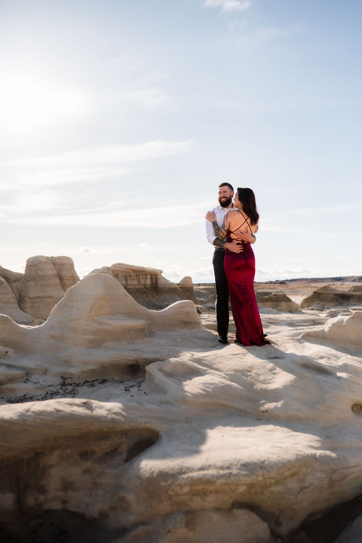 A couple stands intimately on white rock formations against a bright desert sky.