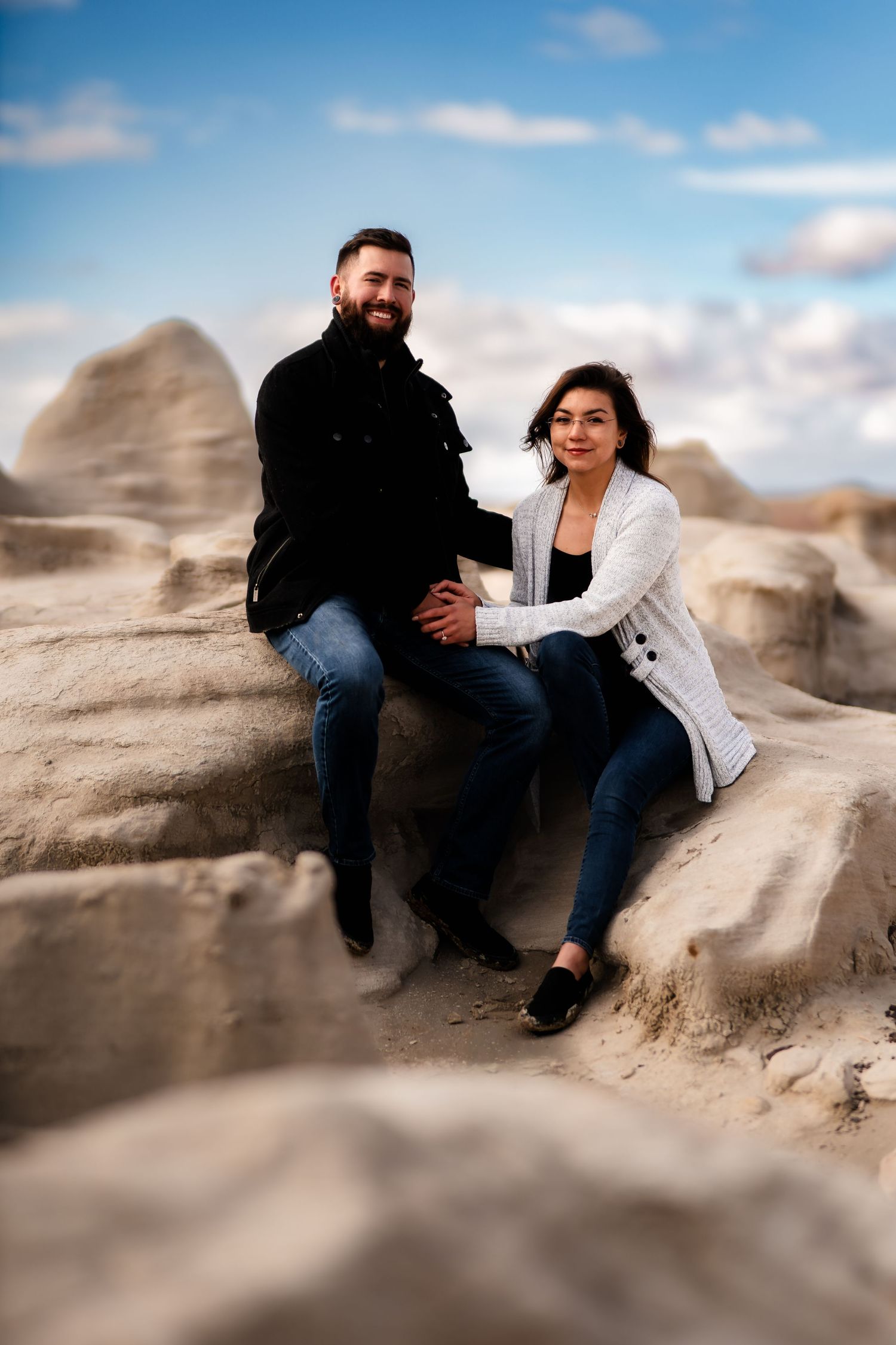 A couple sits together on rugged desert rock formations against a blue sky with white clouds.