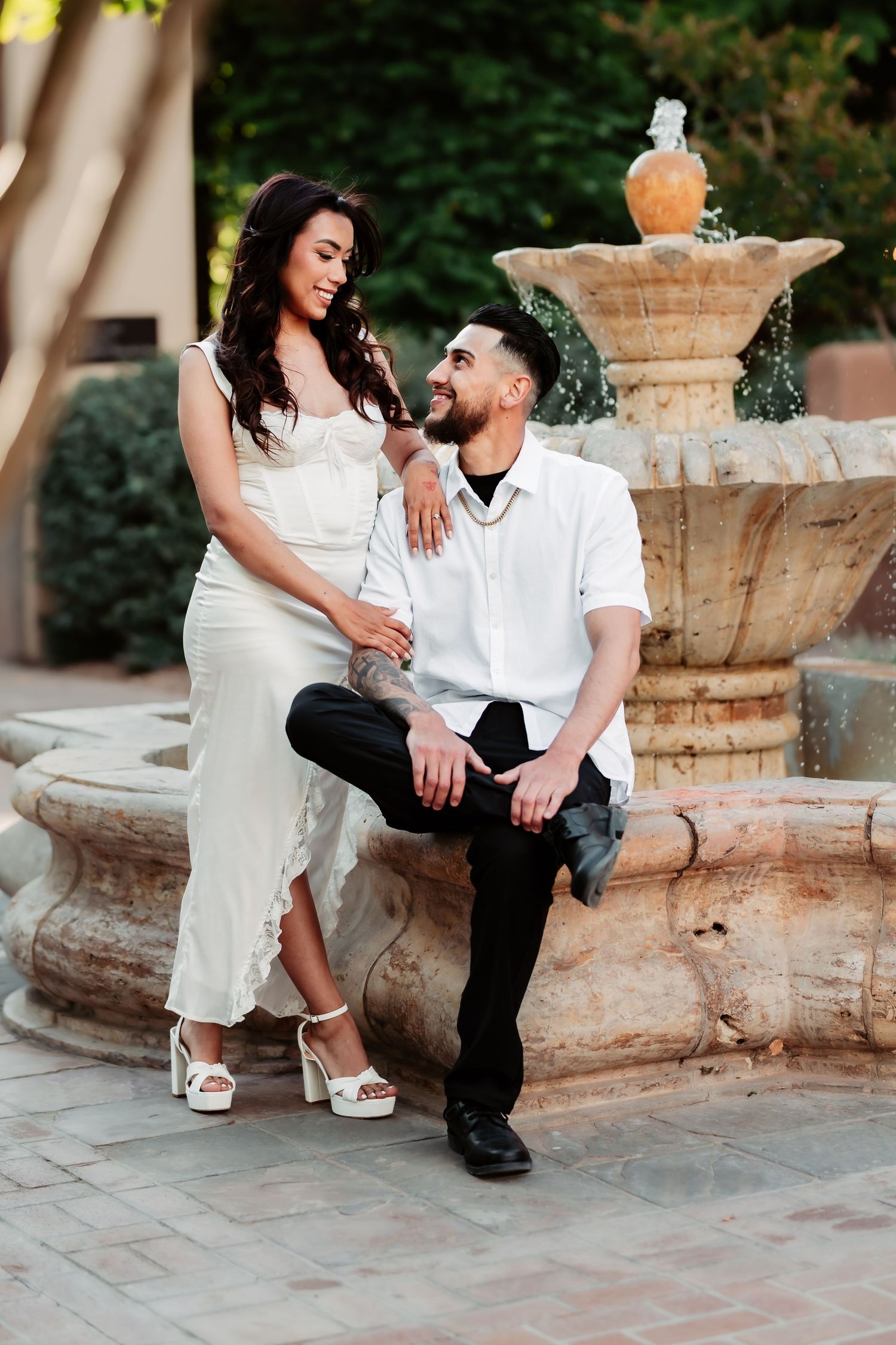 A romantic couple poses together by an ornate stone fountain in matching white outfits.