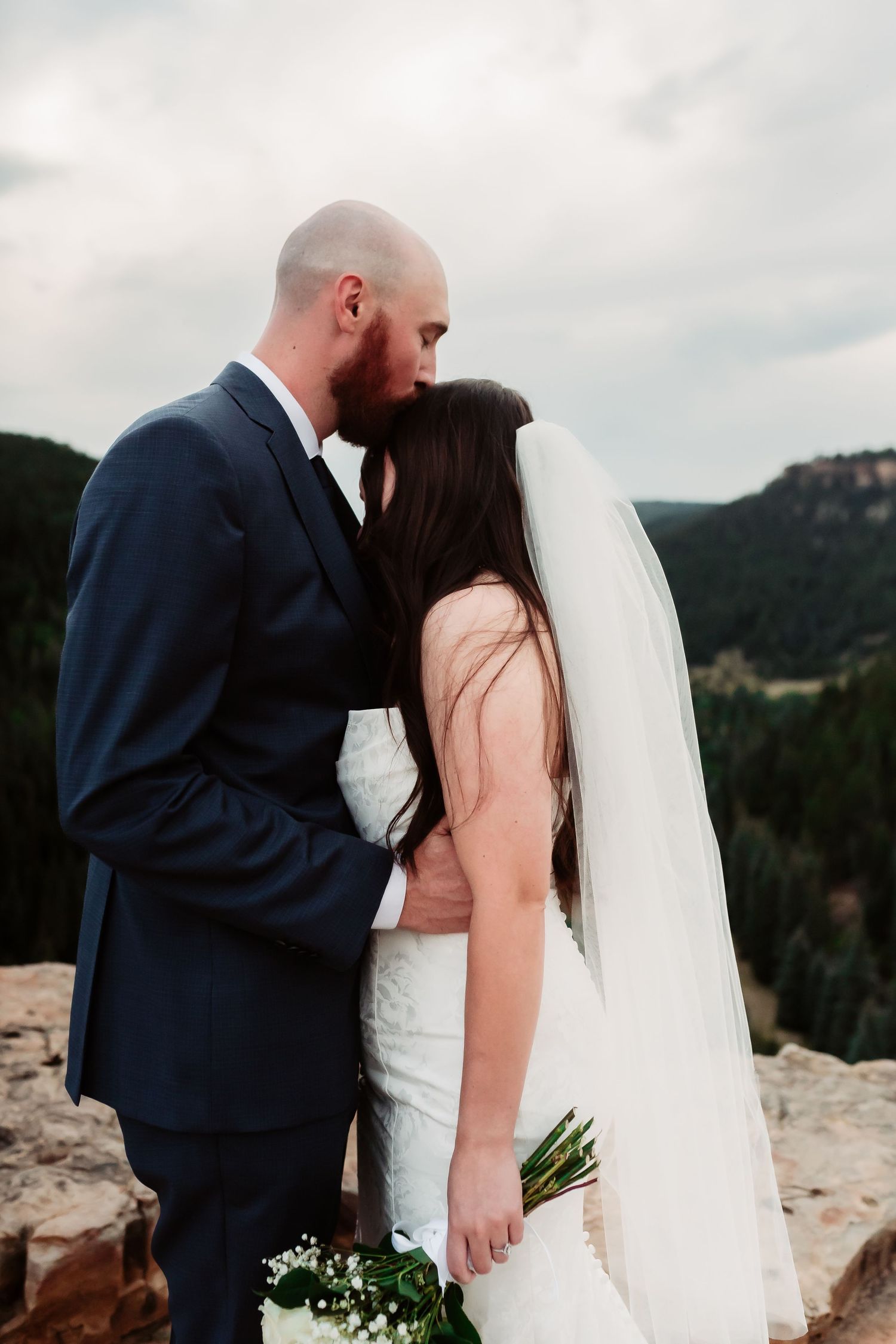 Intimate moment between newlyweds on mountain cliff with scenic forest backdrop.