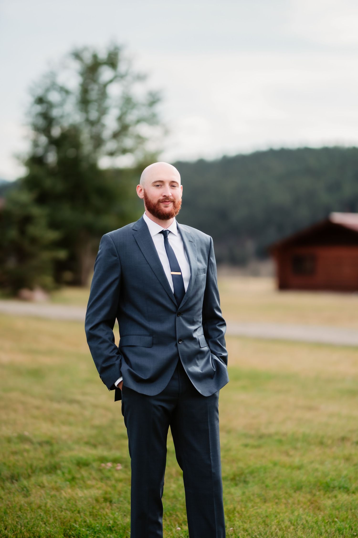 A well-dressed person stands outdoors in a navy suit with mountains and trees in the background.