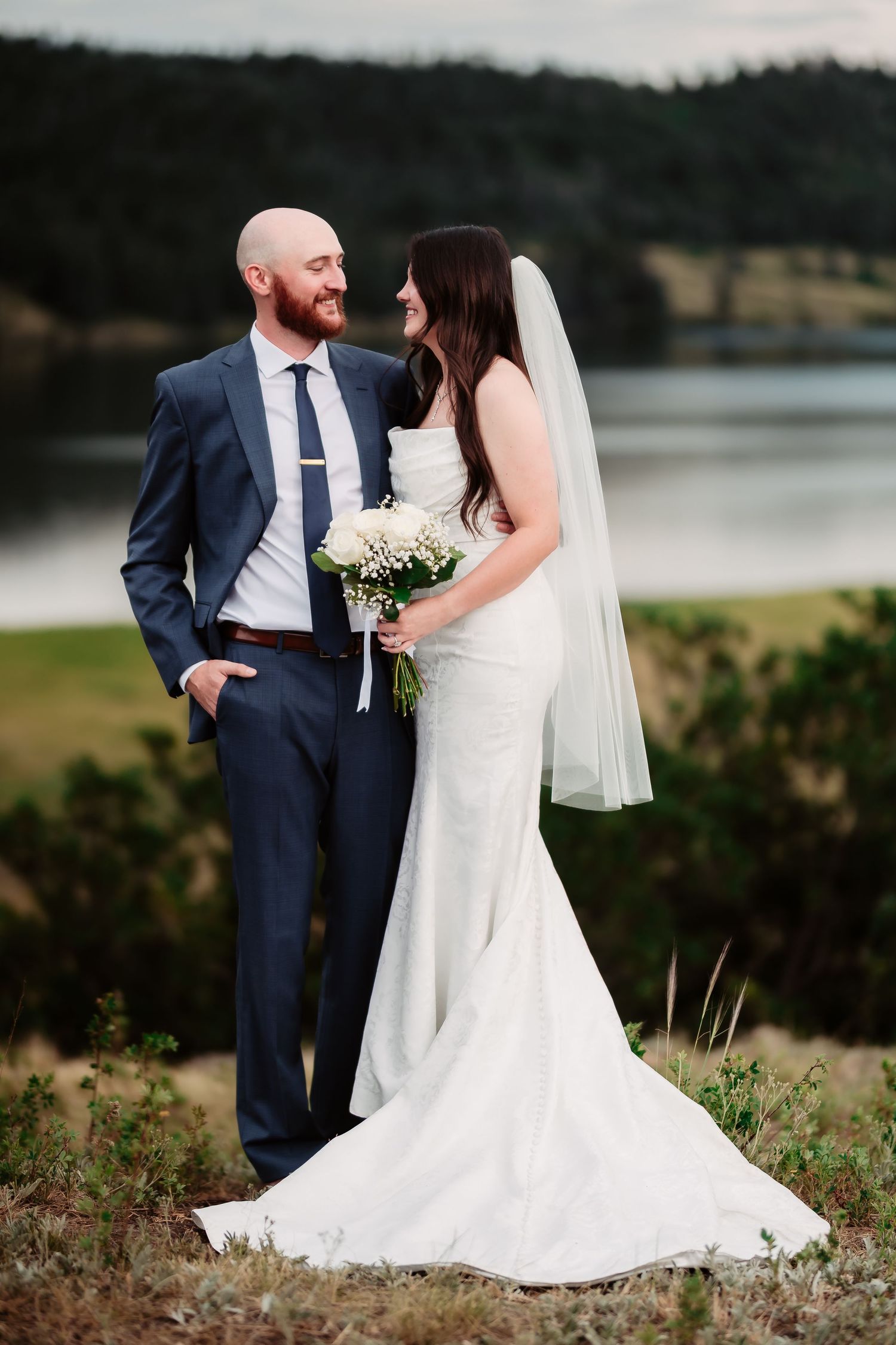 Newlyweds pose together against a lakeside backdrop, bride holding white flower bouquet.