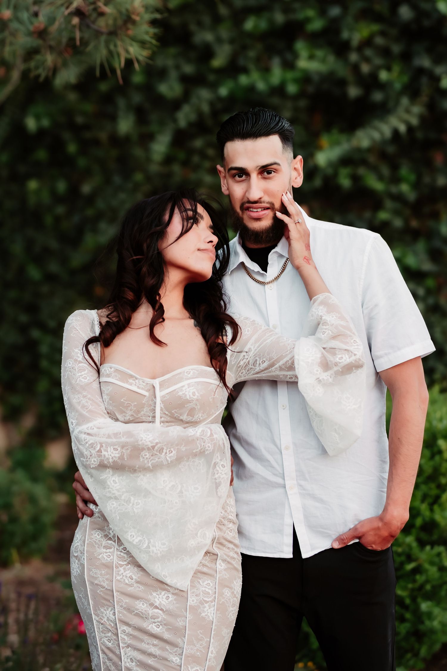 A romantic couple poses outdoors against greenery, with her in a white lace dress and him in a casual white shirt.