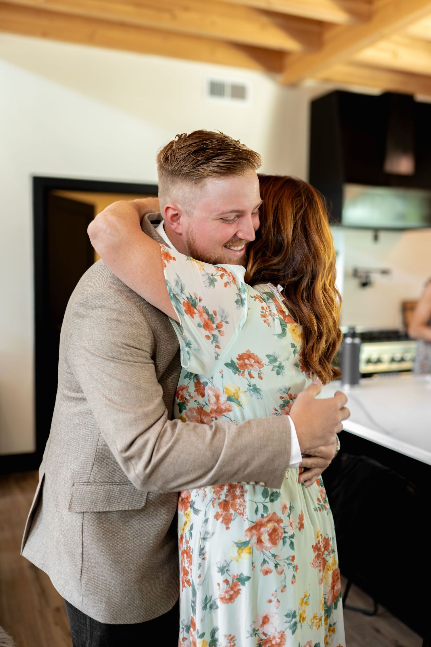 A couple shares a tender embrace in a modern kitchen, with the woman wearing a floral dress.
