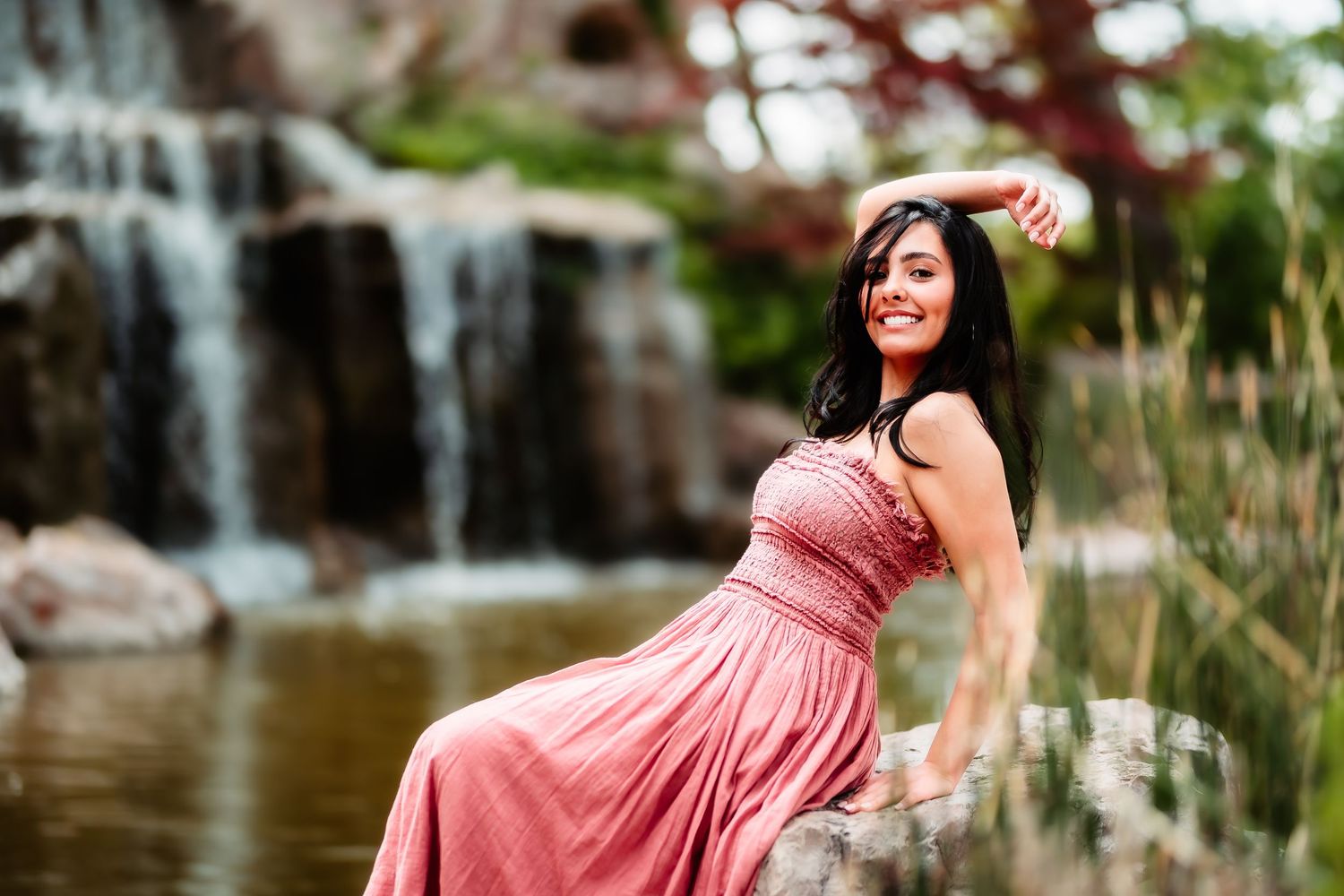 A woman in a flowing pink dress poses elegantly by a scenic waterfall in a garden setting.