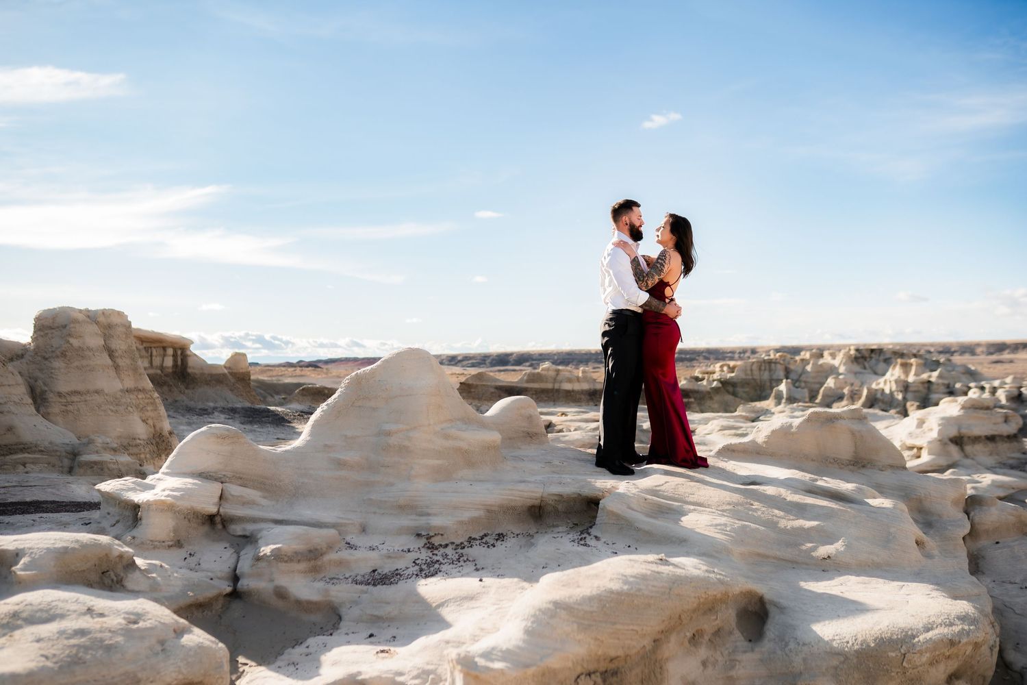 Two people stand together on smooth white rock formations in a desert setting with blue sky overhead.