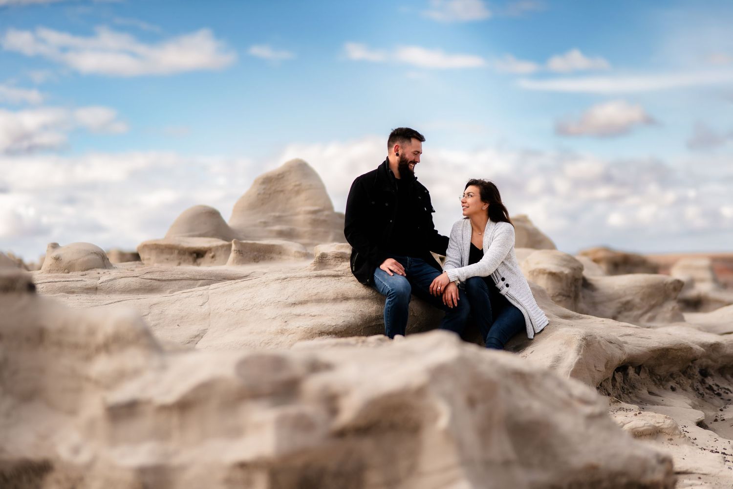 Two people sitting on sandy rock formations under a blue cloudy sky in a desert landscape.