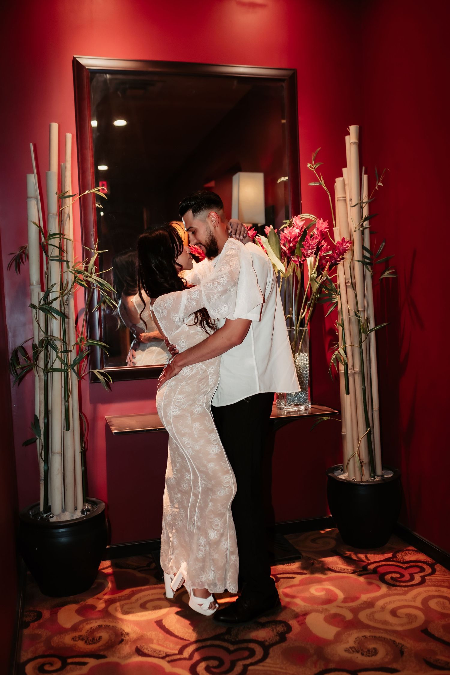 Romantic couple embracing in a red-lit room decorated with bamboo stalks and a mirror.