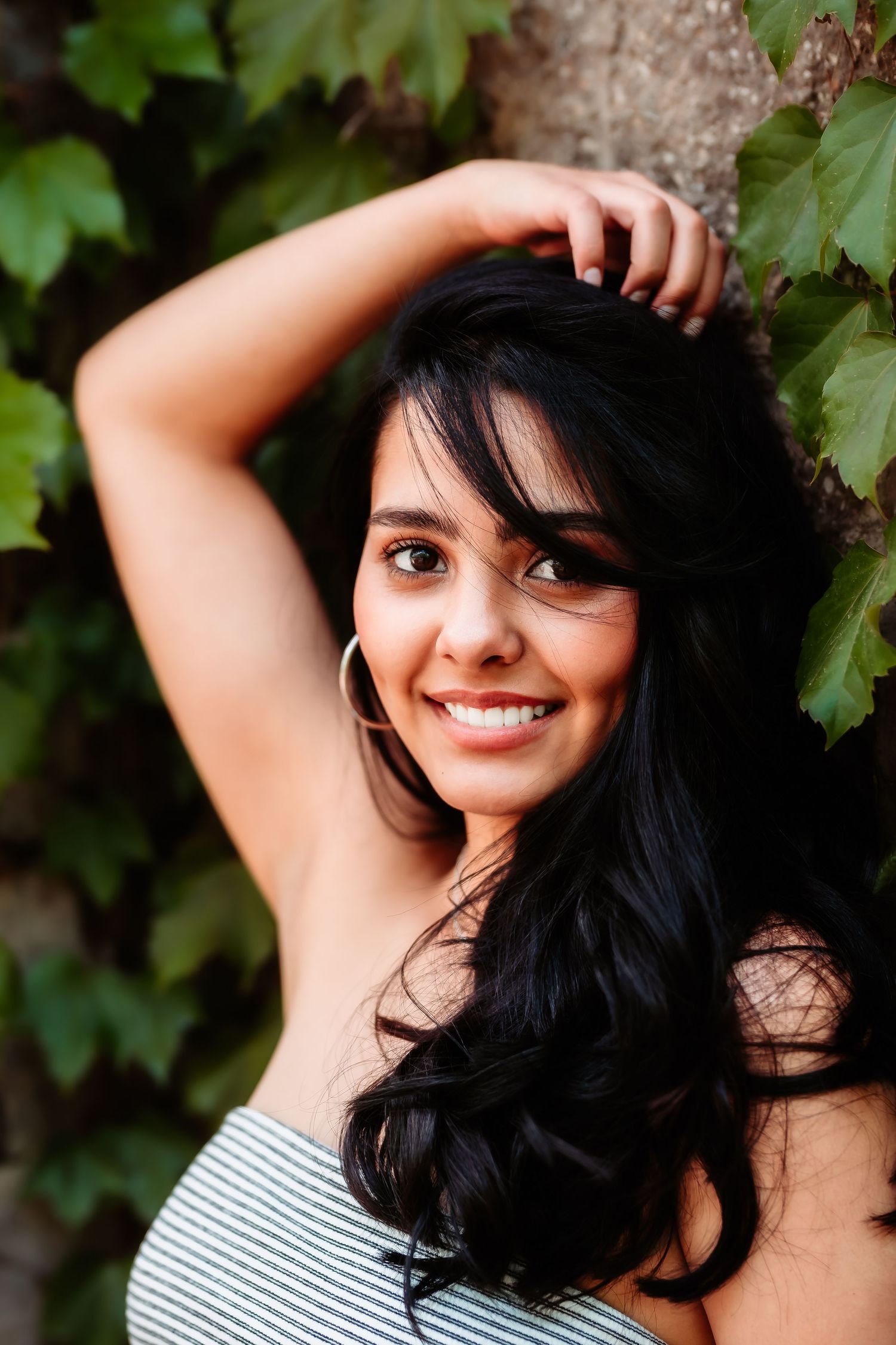 Portrait against ivy-covered wall showing long dark hair and striped top in natural lighting.