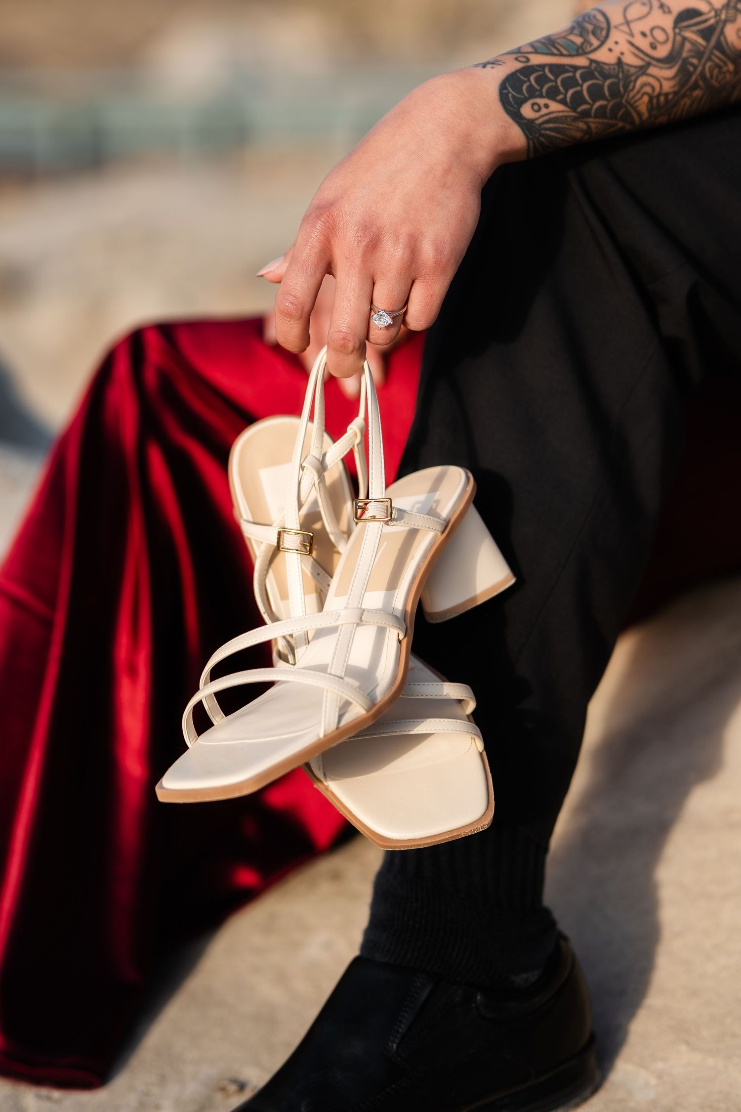 A tattooed hand holds elegant white sandals against a backdrop of red velvet fabric.