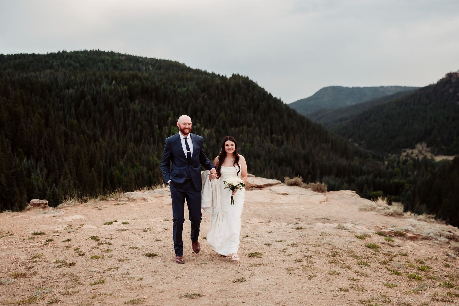 Scenic wedding photos of couple walking along mountain ridge with forest valley views.