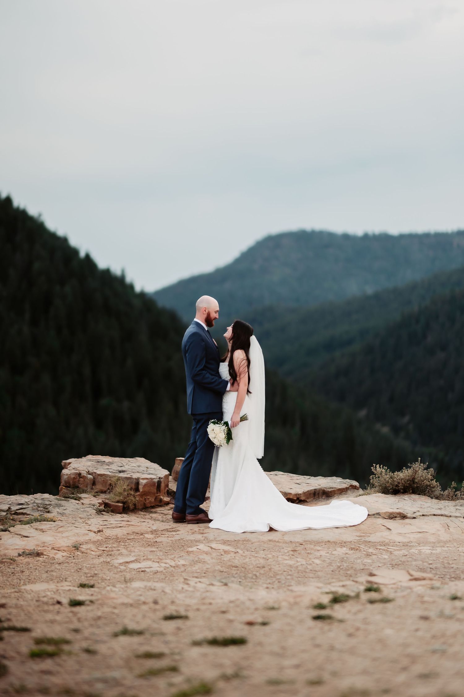 Newlyweds embrace on a mountain cliff edge with a dramatic valley vista behind them.