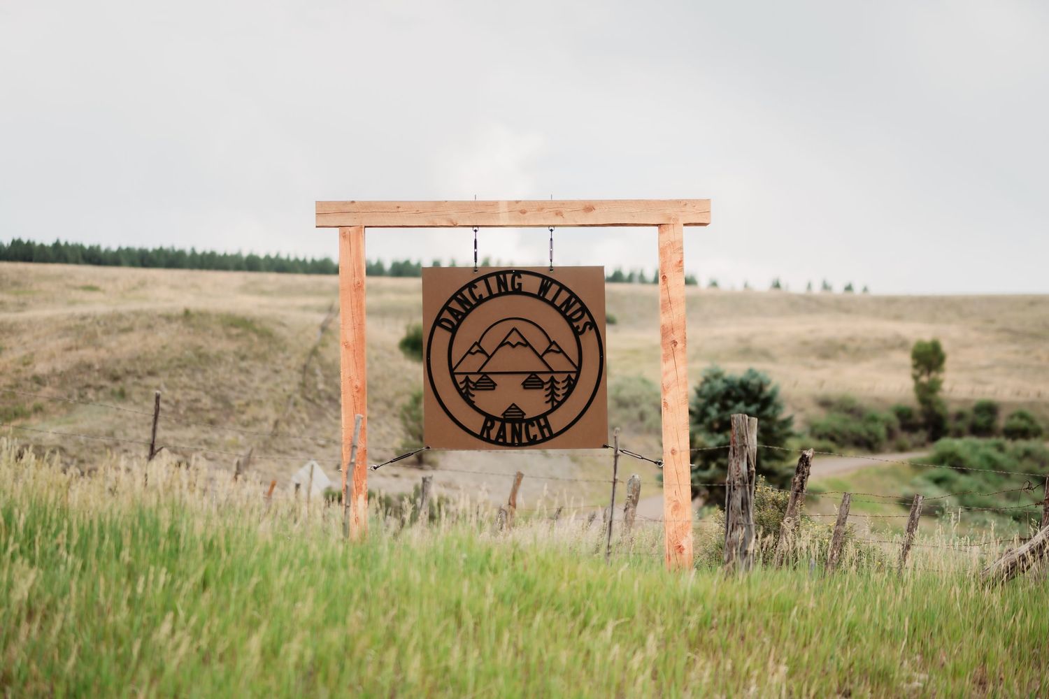 Wooden entrance sign with mountain logo framed by orange posts in a grassy field landscape.