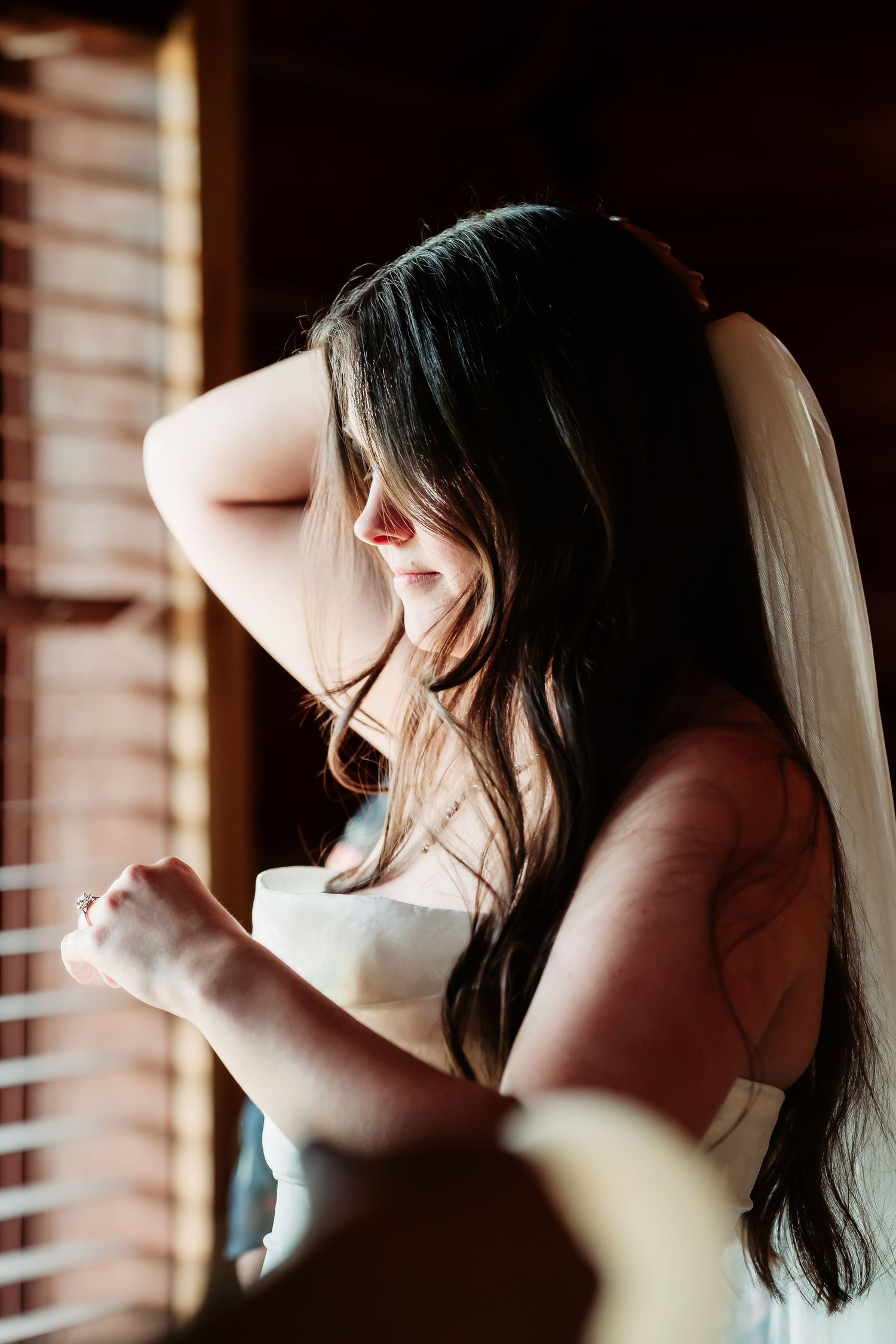 Sunlit silhouette of someone in a wedding dress getting ready by a window with soft natural light.