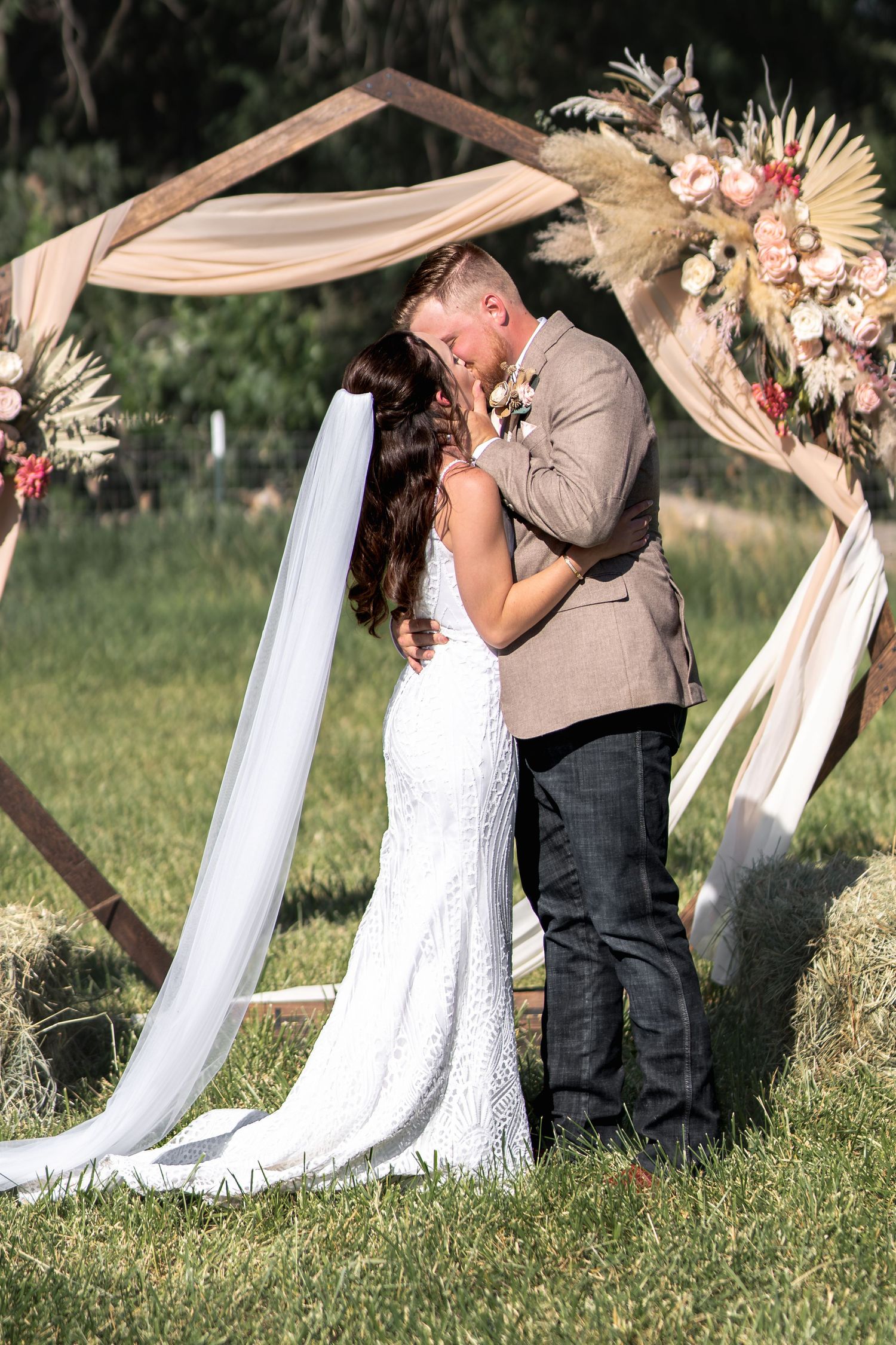 A romantic wedding ceremony under a draped arch decorated with dried flowers and pampas grass in an outdoor garden setting.