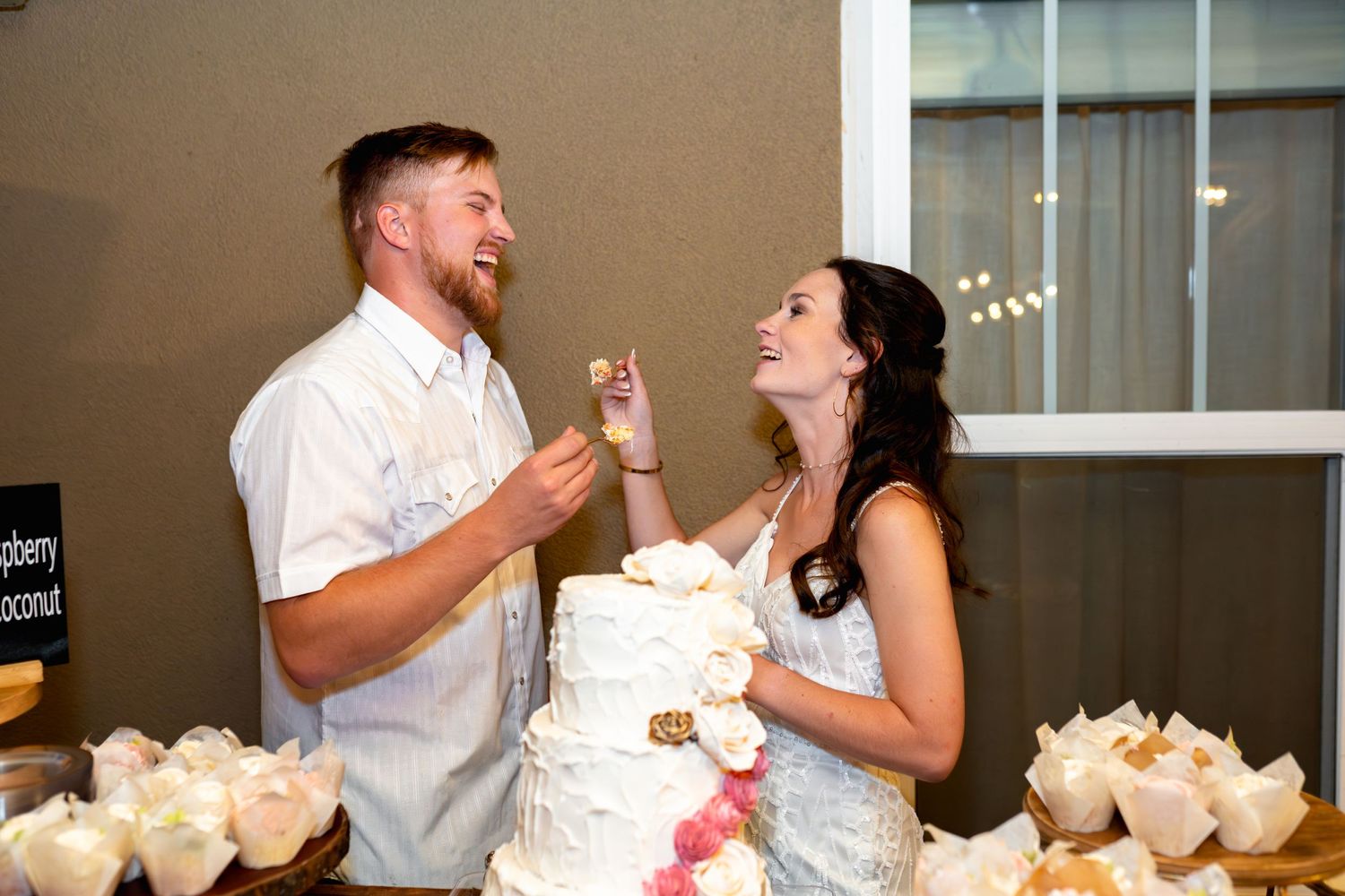 Happy couple playfully shares wedding cake at their reception while standing next to white dessert table.