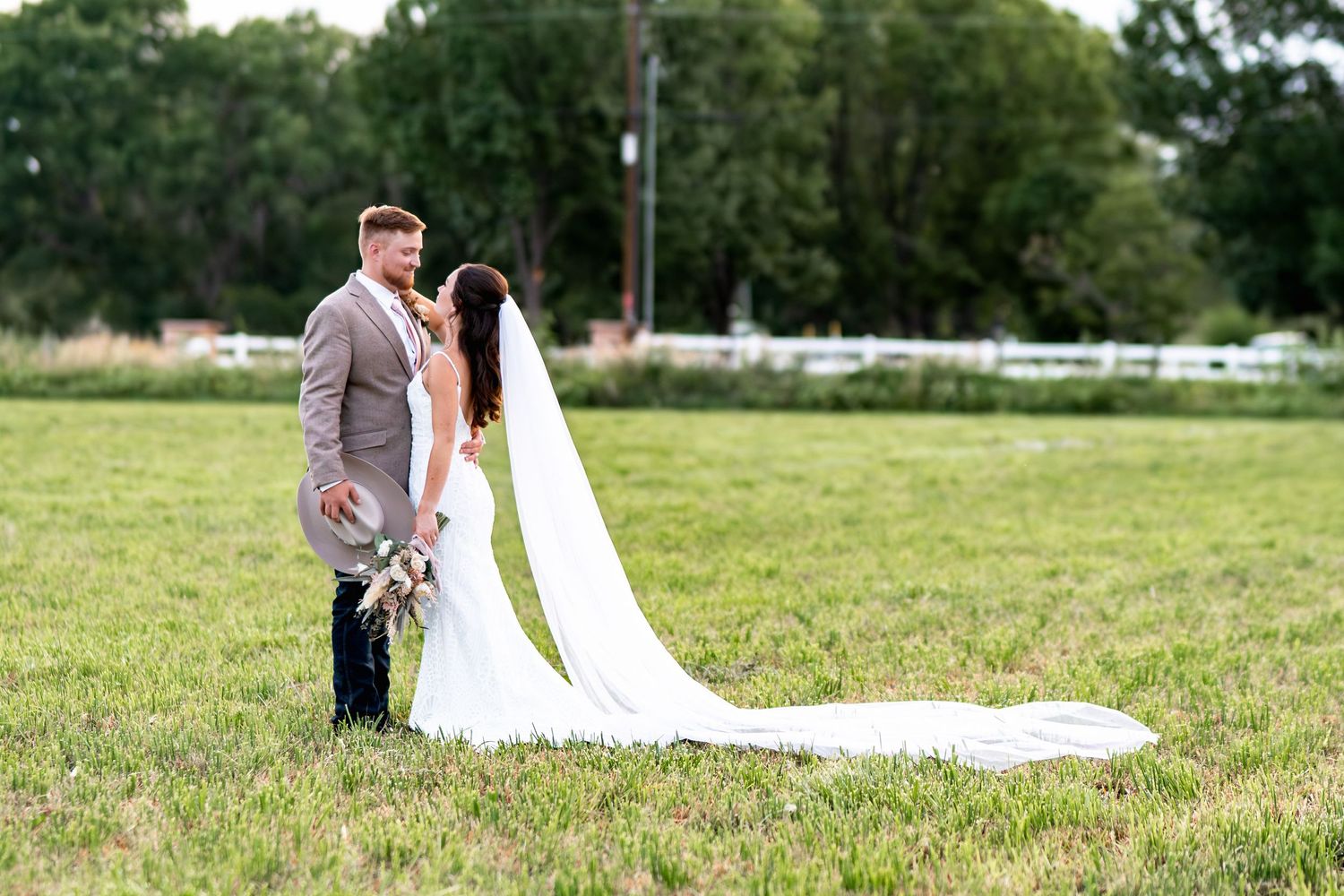 A couple shares a romantic moment in their wedding attire on a grassy field with her long white veil flowing.