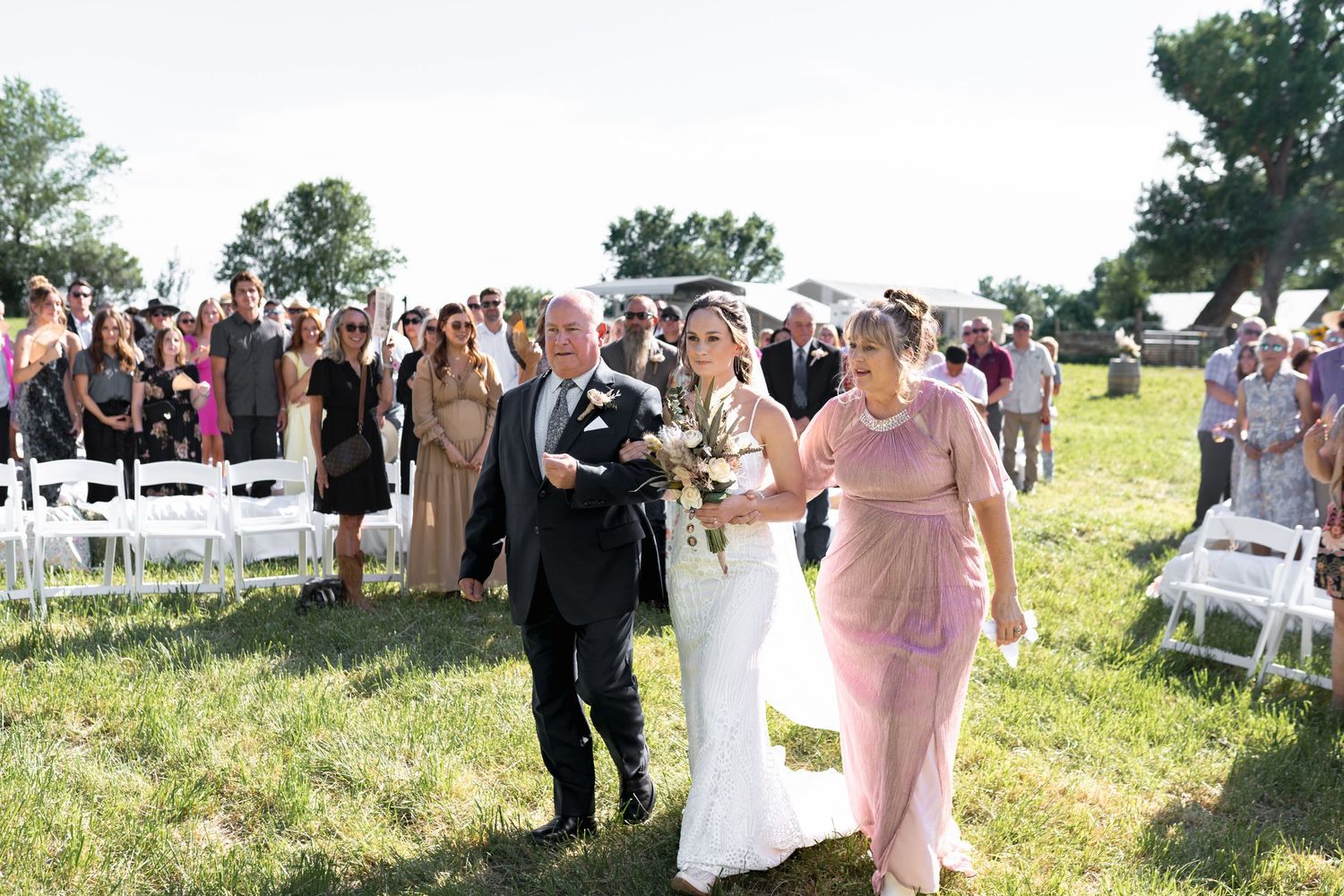 Wedding party members walk down a grassy aisle during an outdoor ceremony with white chairs and guests.