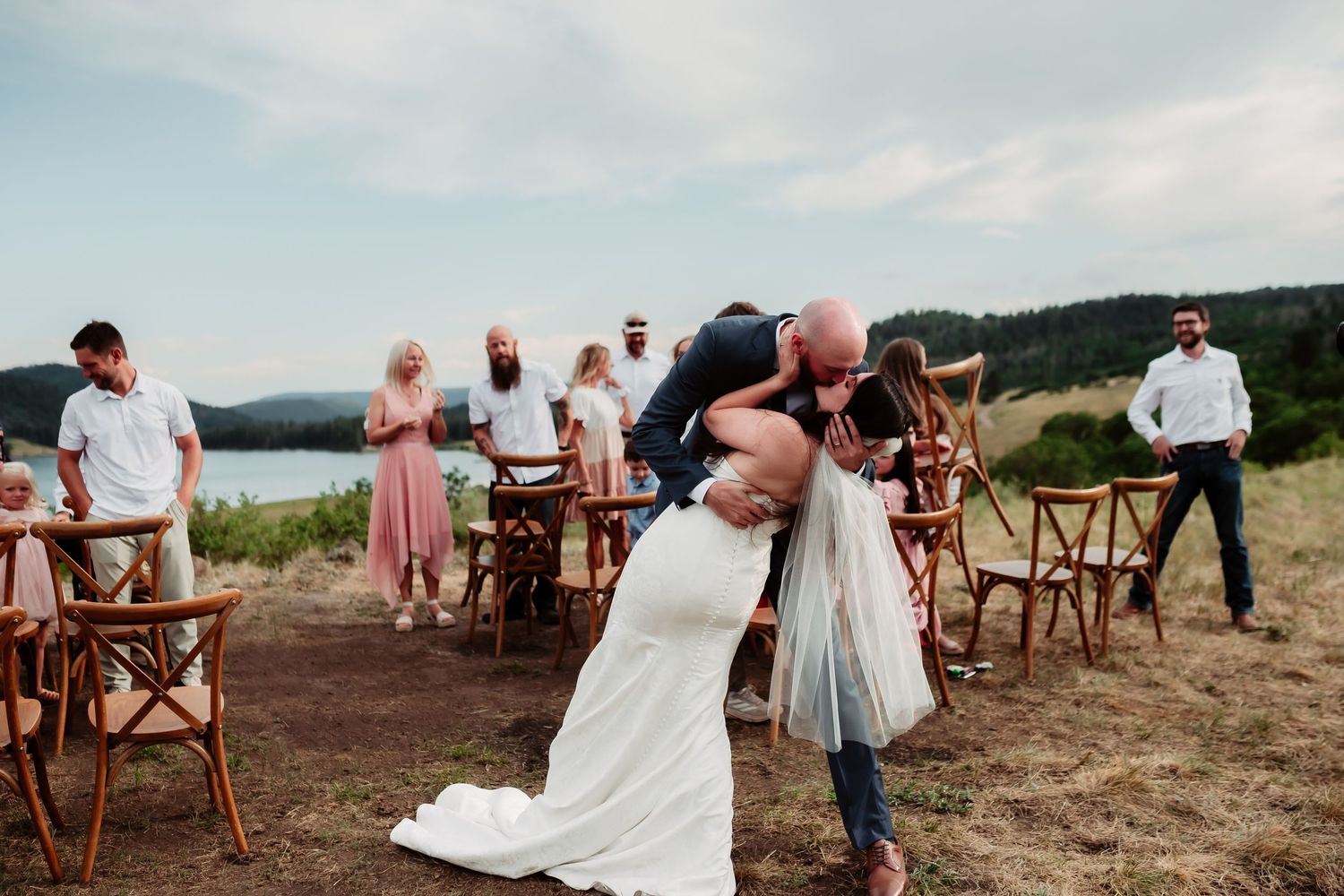 A romantic first kiss at an outdoor wedding with guests and wooden chairs surrounding the couple.