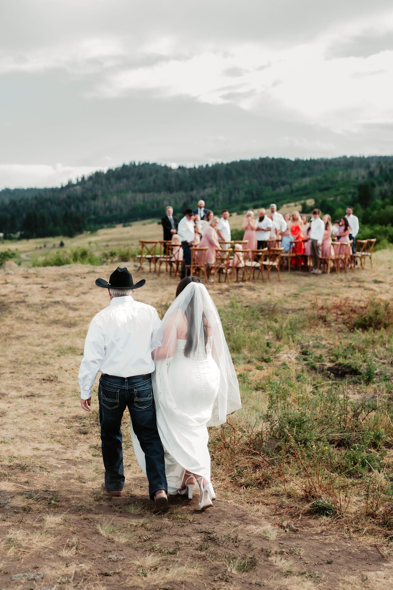 Outdoor wedding ceremony on a hilltop with wooden chairs and couple walking down dirt path.