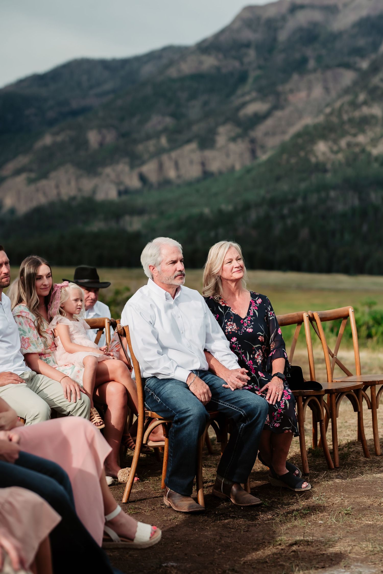 Wedding guests watch ceremony against dramatic mountain backdrop with wooden chairs.