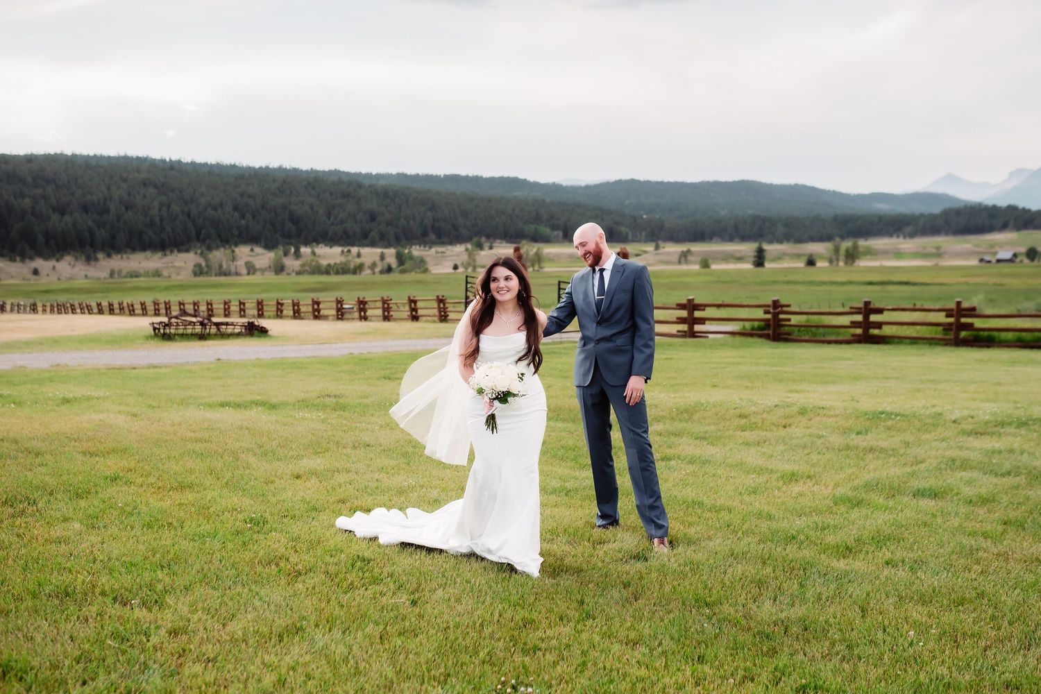 Panoramic view of a wedding couple walking through a grassy field with mountain vistas.