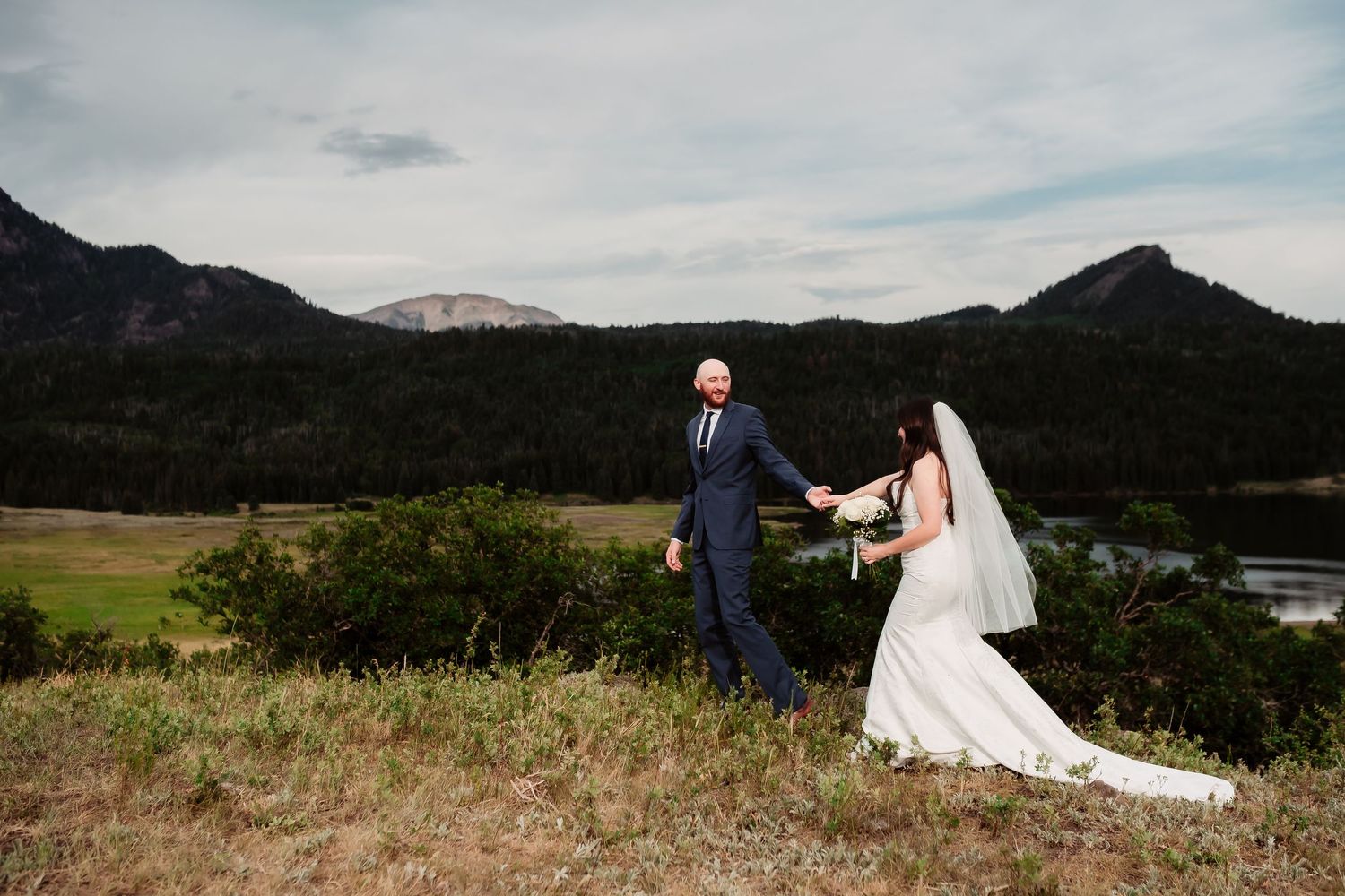 Couple walking hand in hand on a hilltop with mountain views and lake in background.