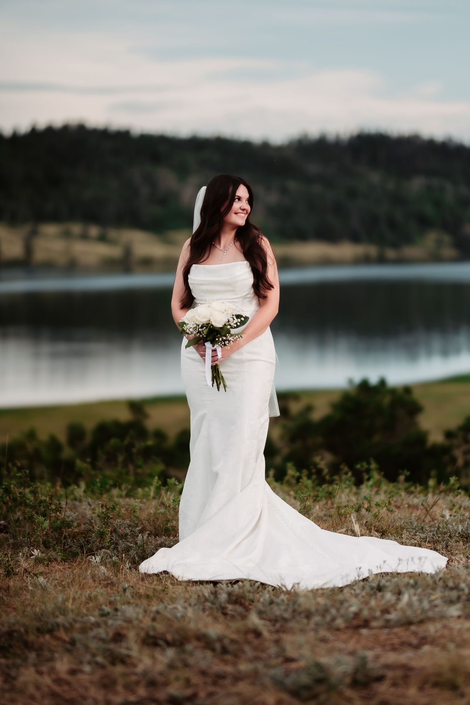 A bride in a fitted white gown holds a bouquet while posing by a serene lake at sunset.