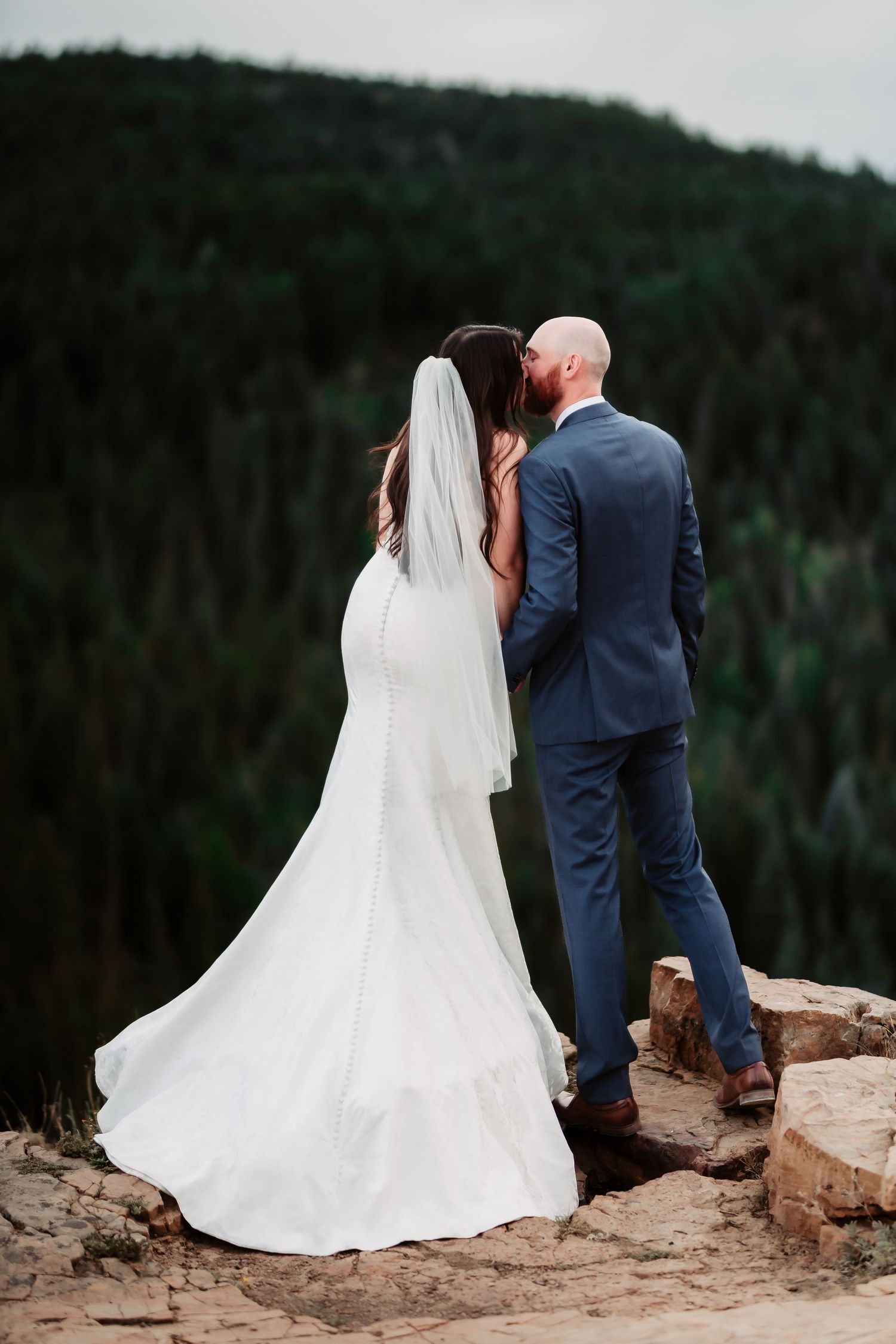 Elegant wedding couple standing on cliff edge overlooking forested mountains with bride in flowing white gown.