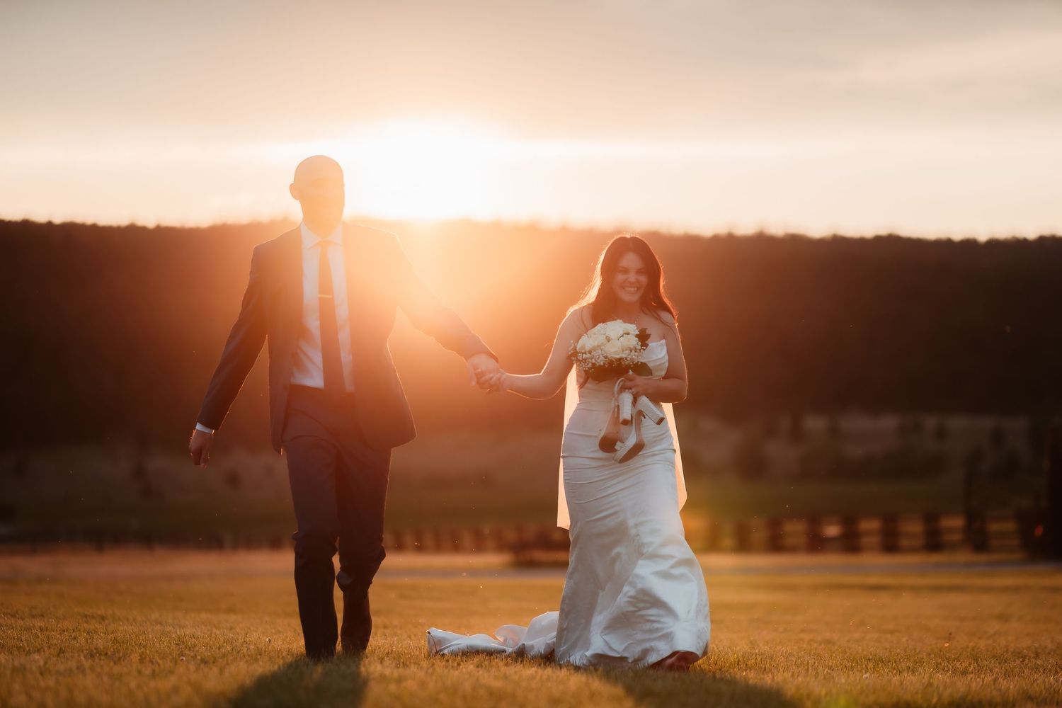 A couple walks hand in hand during sunset in their wedding attire across an open field.