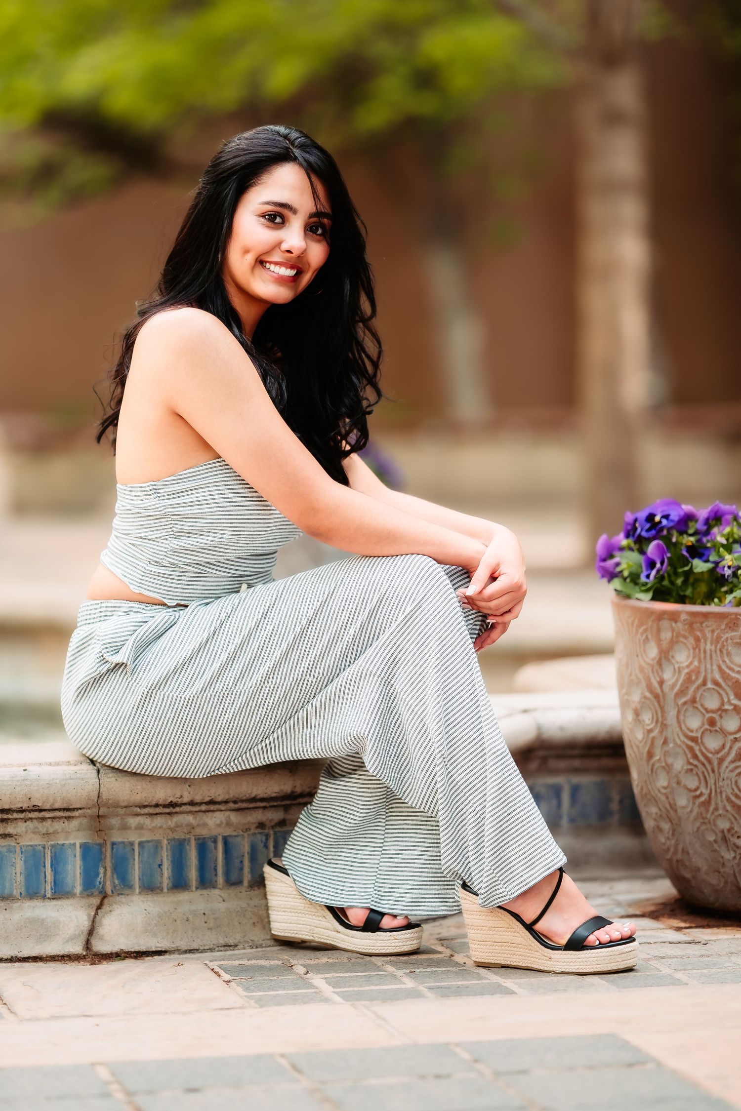 A casual portrait of a woman in gray loungewear sitting on stone steps near purple flowers.
