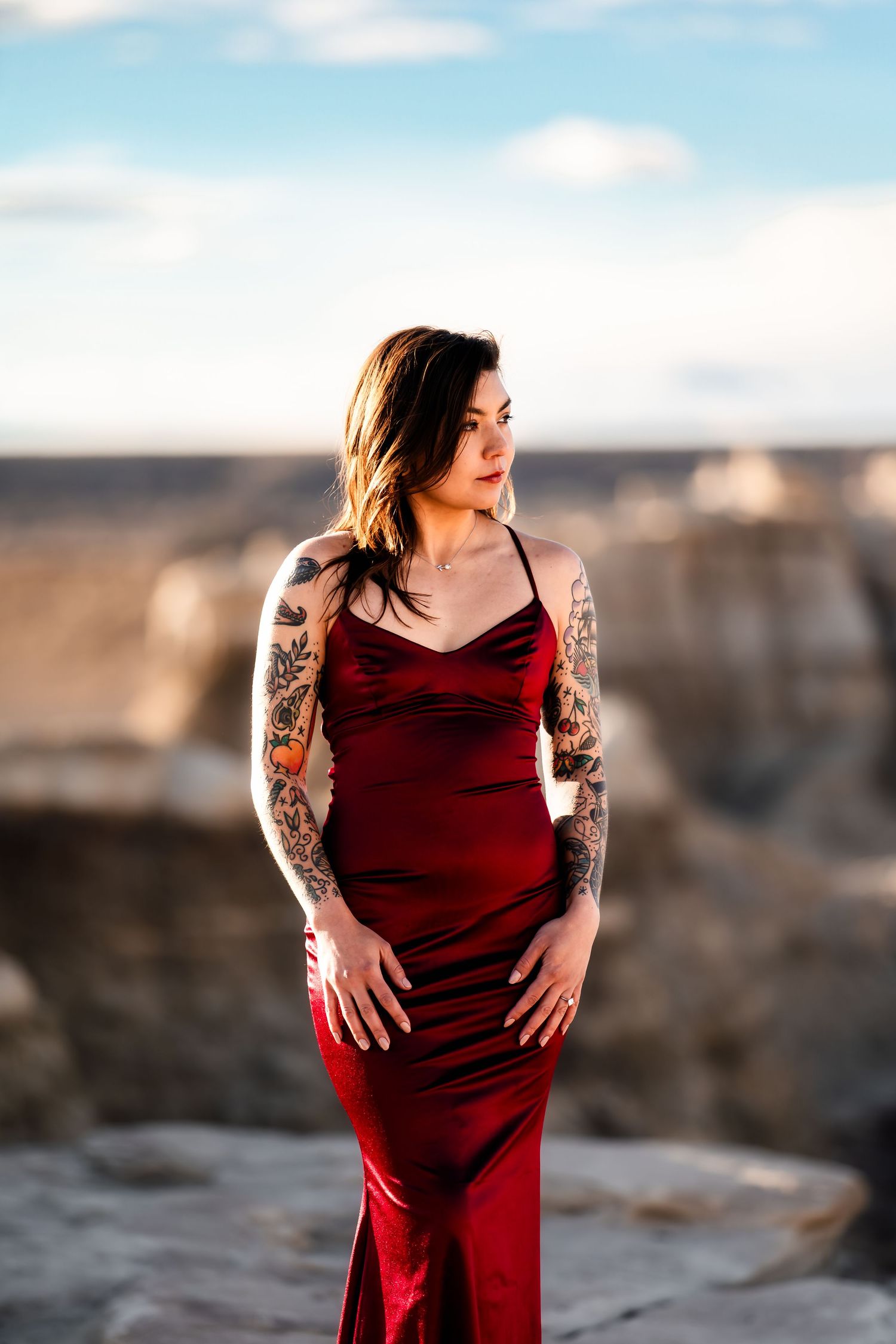 Woman in red gown with sleeve tattoos poses on rocky desert landscape.