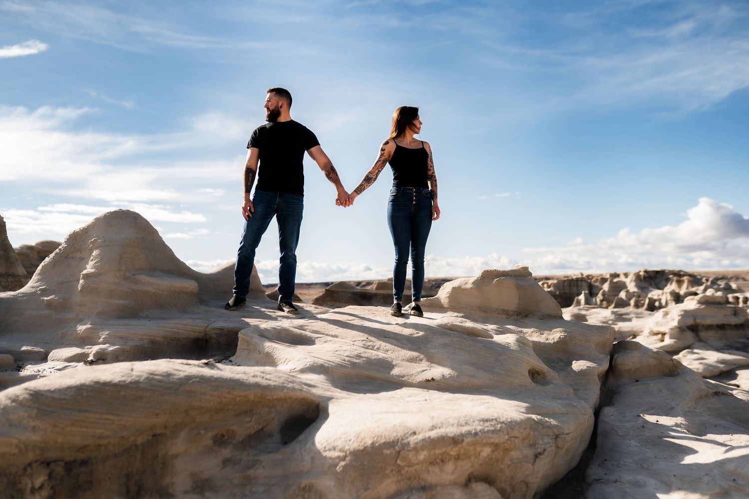 Two people hold hands while standing on smooth white rock formations against a bright blue desert sky.