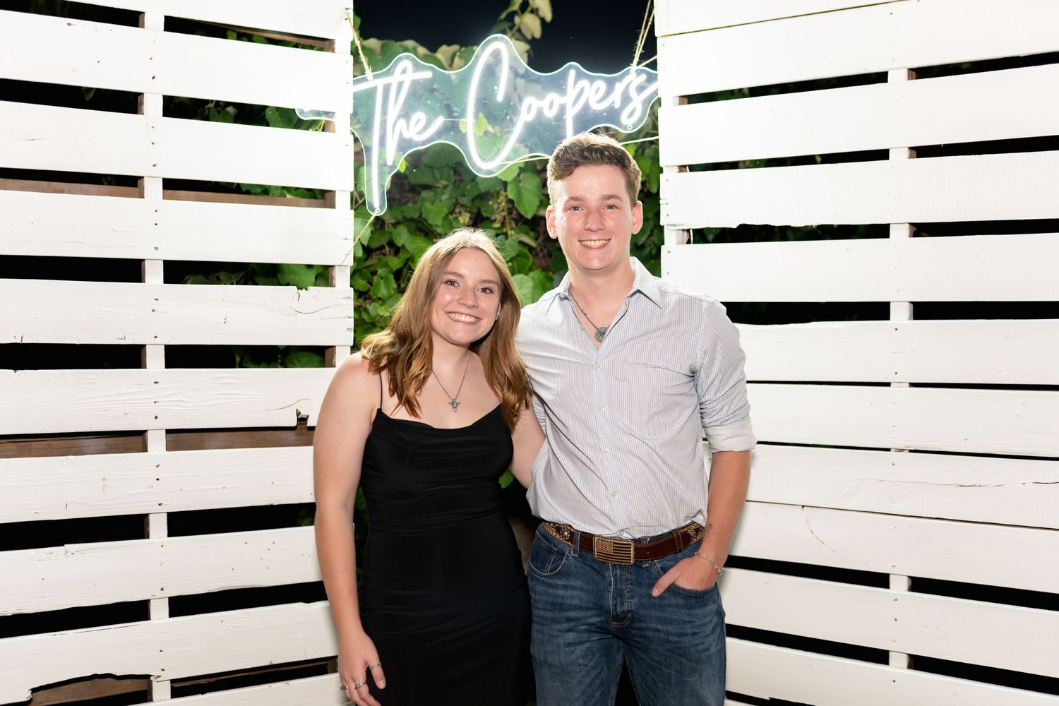 A couple poses in front of a white slatted wall backdrop with neon signage at an event.