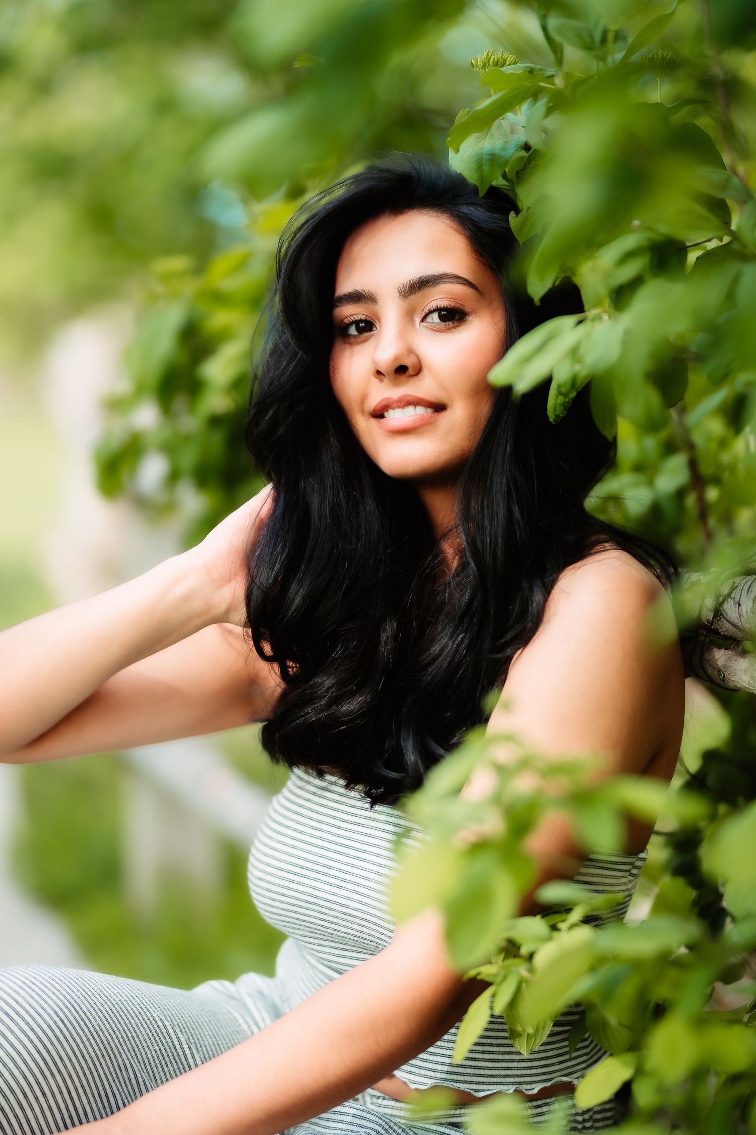 A candid outdoor portrait among green foliage wearing a striped top with flowing dark hair.