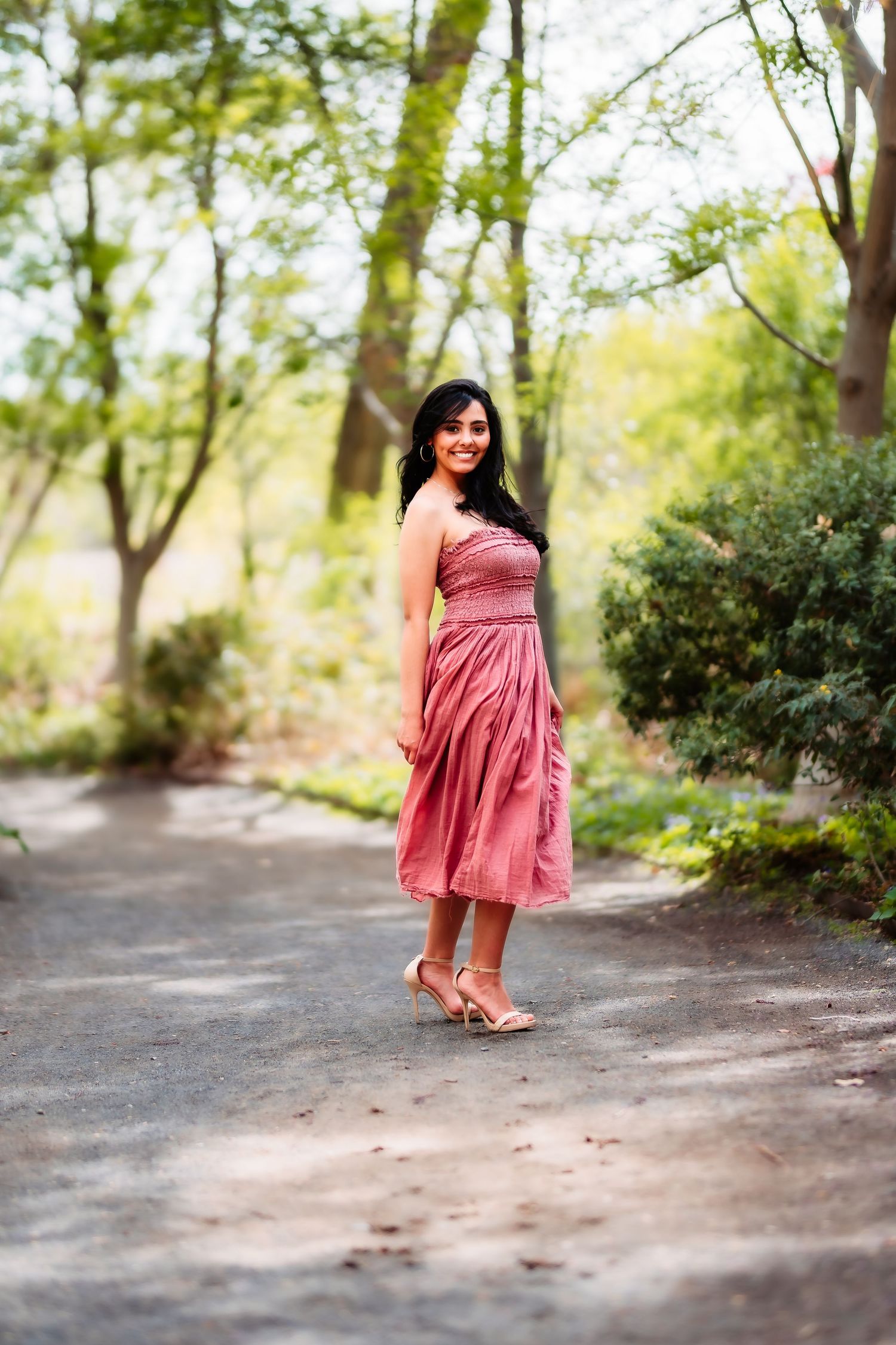 Woman in a pink dress standing on a garden path surrounded by lush green trees and foliage.