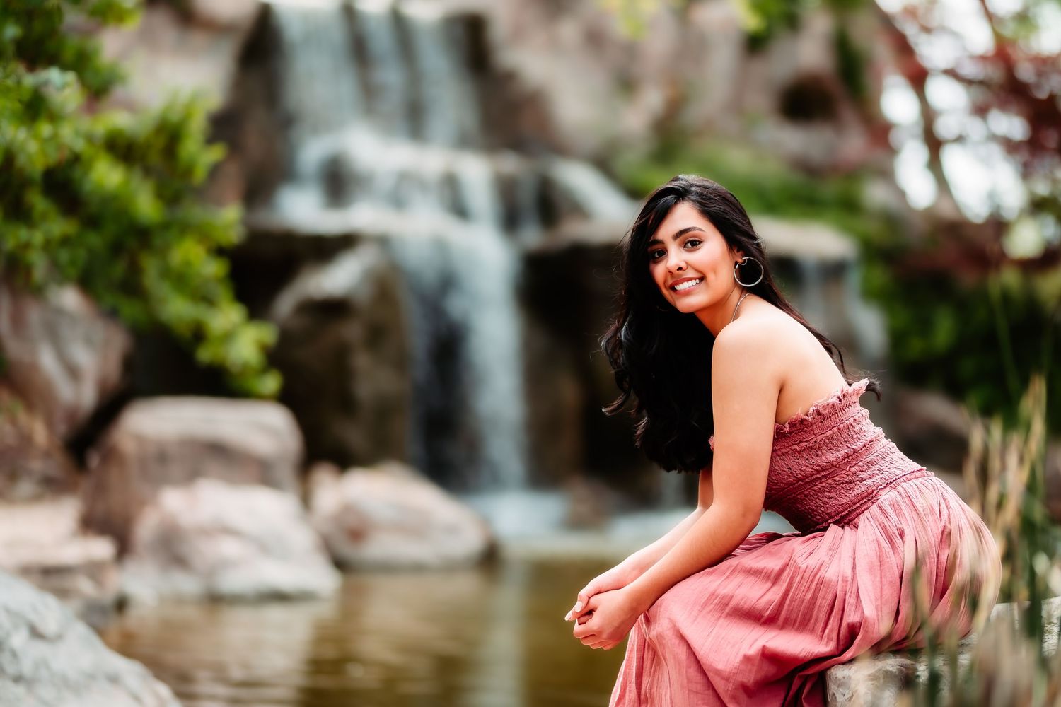 Woman in a flowing pink dress poses beside a scenic waterfall and pond in a garden setting.