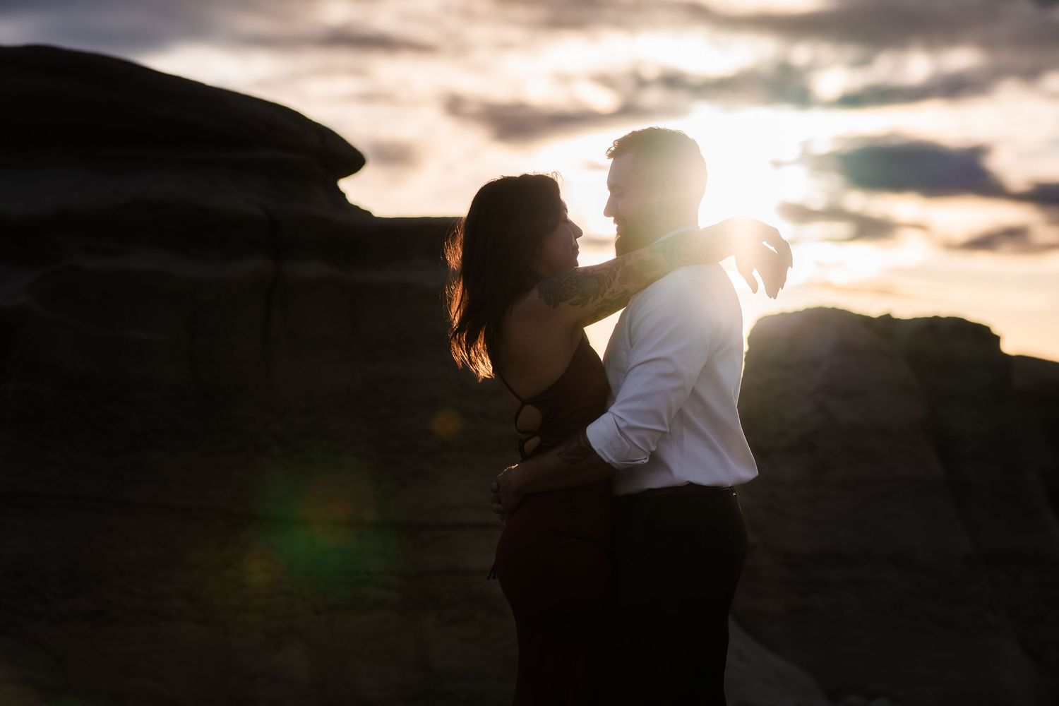 Silhouetted couple embracing against dramatic sunset sky with rock formations in background and lens flare.