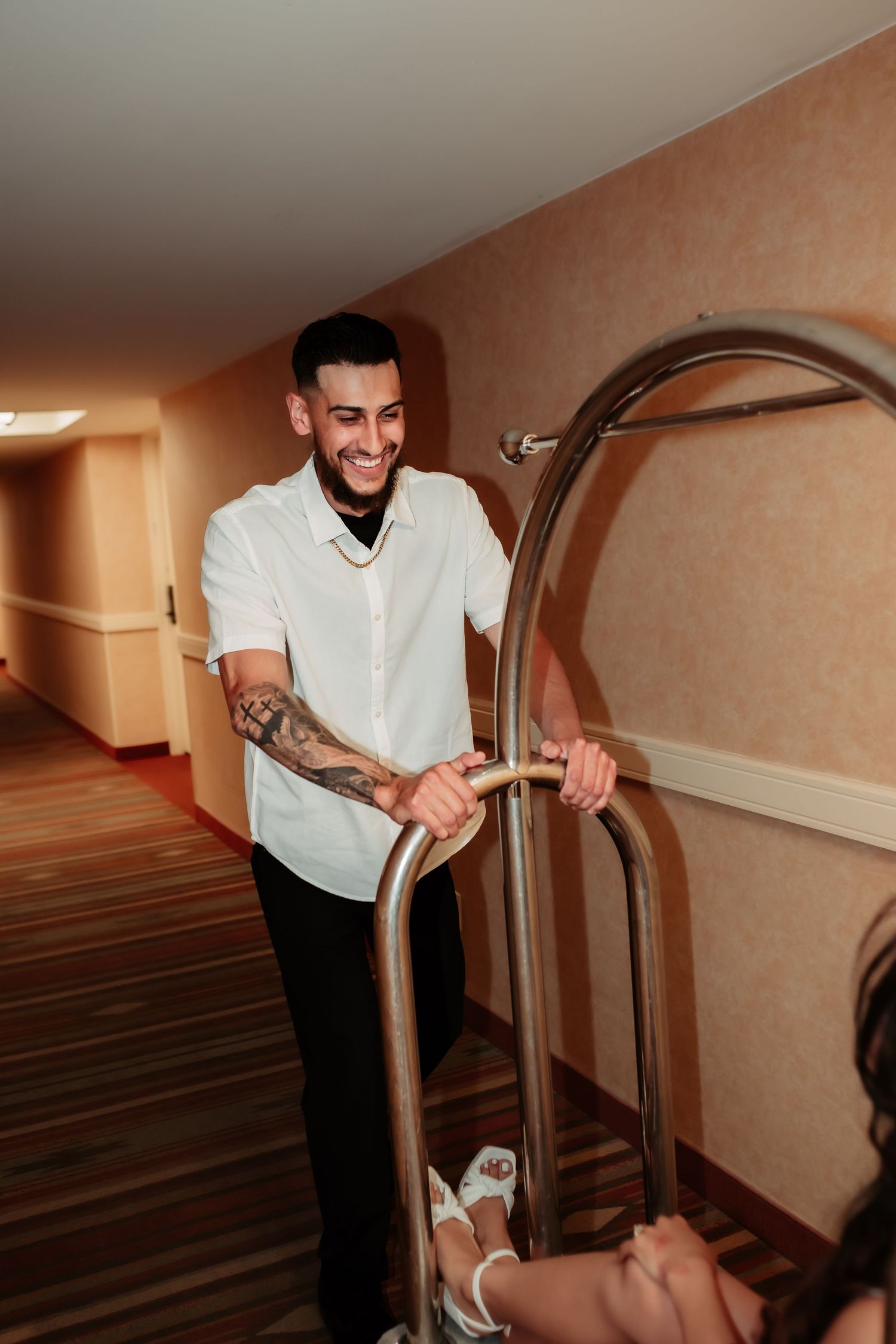 Man in white shirt smiles while pushing a luggage cart in hotel hallway.