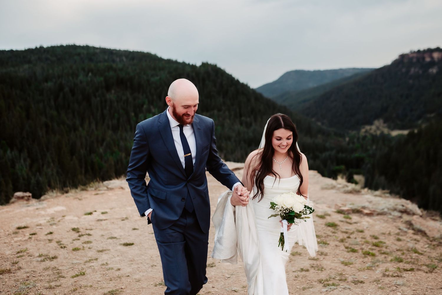 A joyful wedding couple walking together on a mountain summit with evergreen forests below.