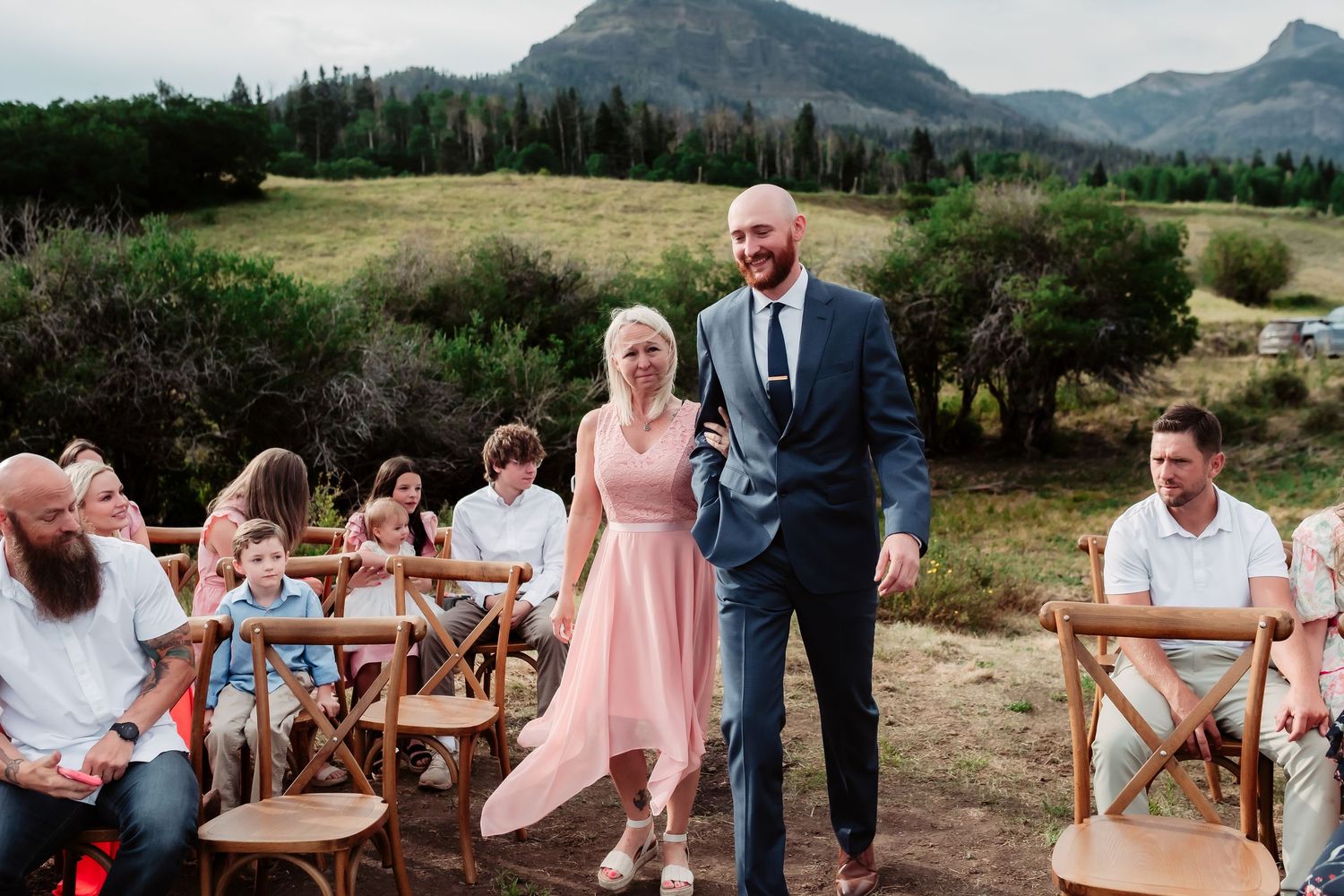 Wedding guests seated at wooden chairs during mountain ceremony with scenic backdrop.
